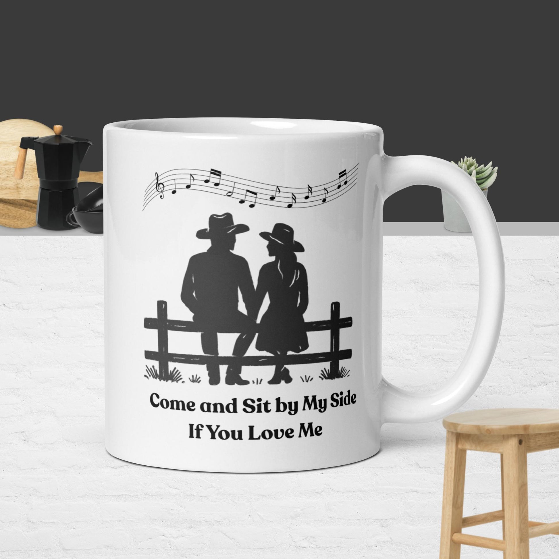 White mug with a design of two cowboy lovers sitting on a fence and text, held by a hand on a wooden table.