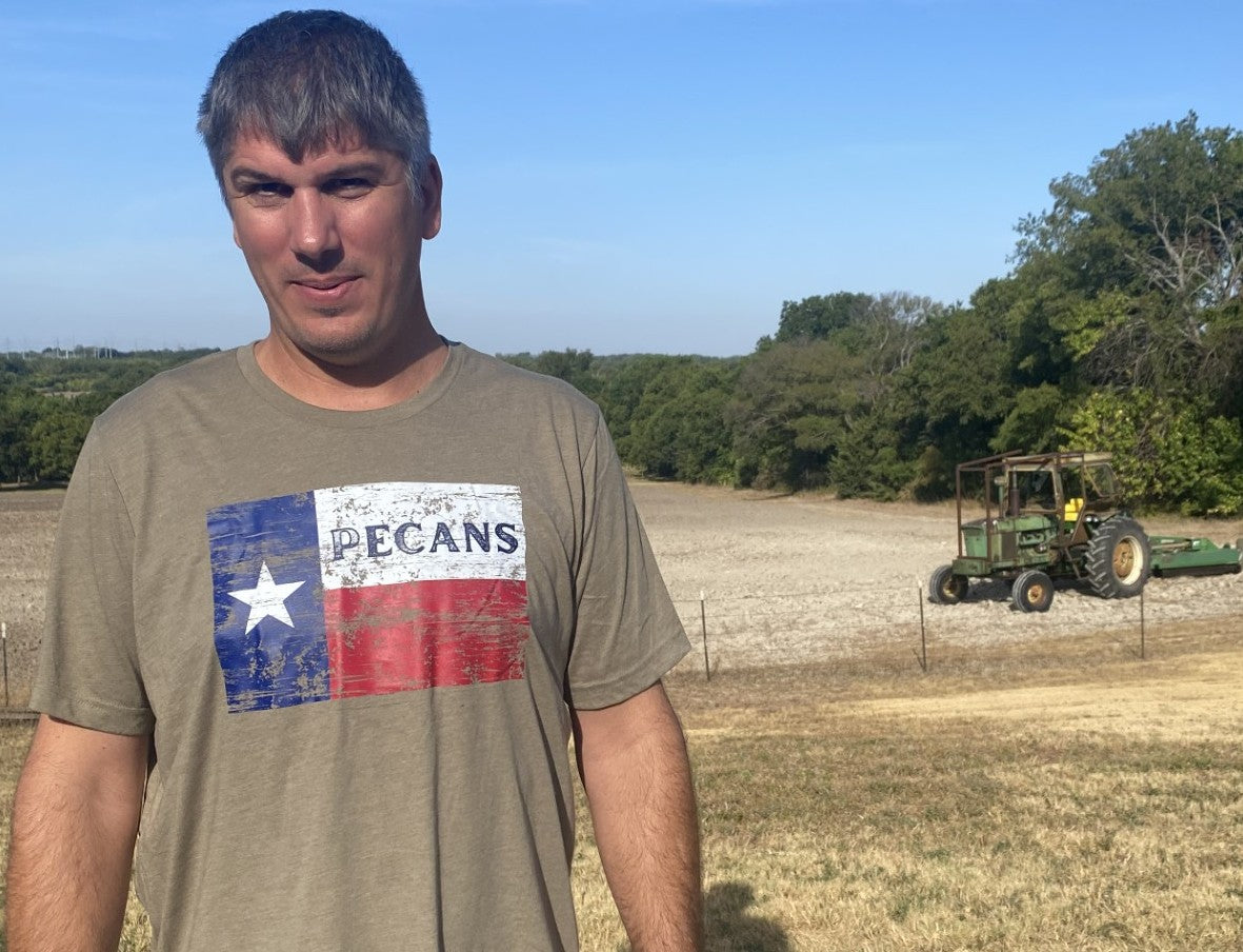 A man wearing a tan t-shirt with a distressed Texas flag graphic that reads 'PECANS' stands in a field, with a tractor working in the background under a blue sky.