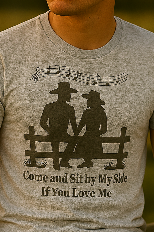Young cowboy in heather gray tee sitting on a fence, looking wistfully into the sunset with heart-shaped bokeh in the background.