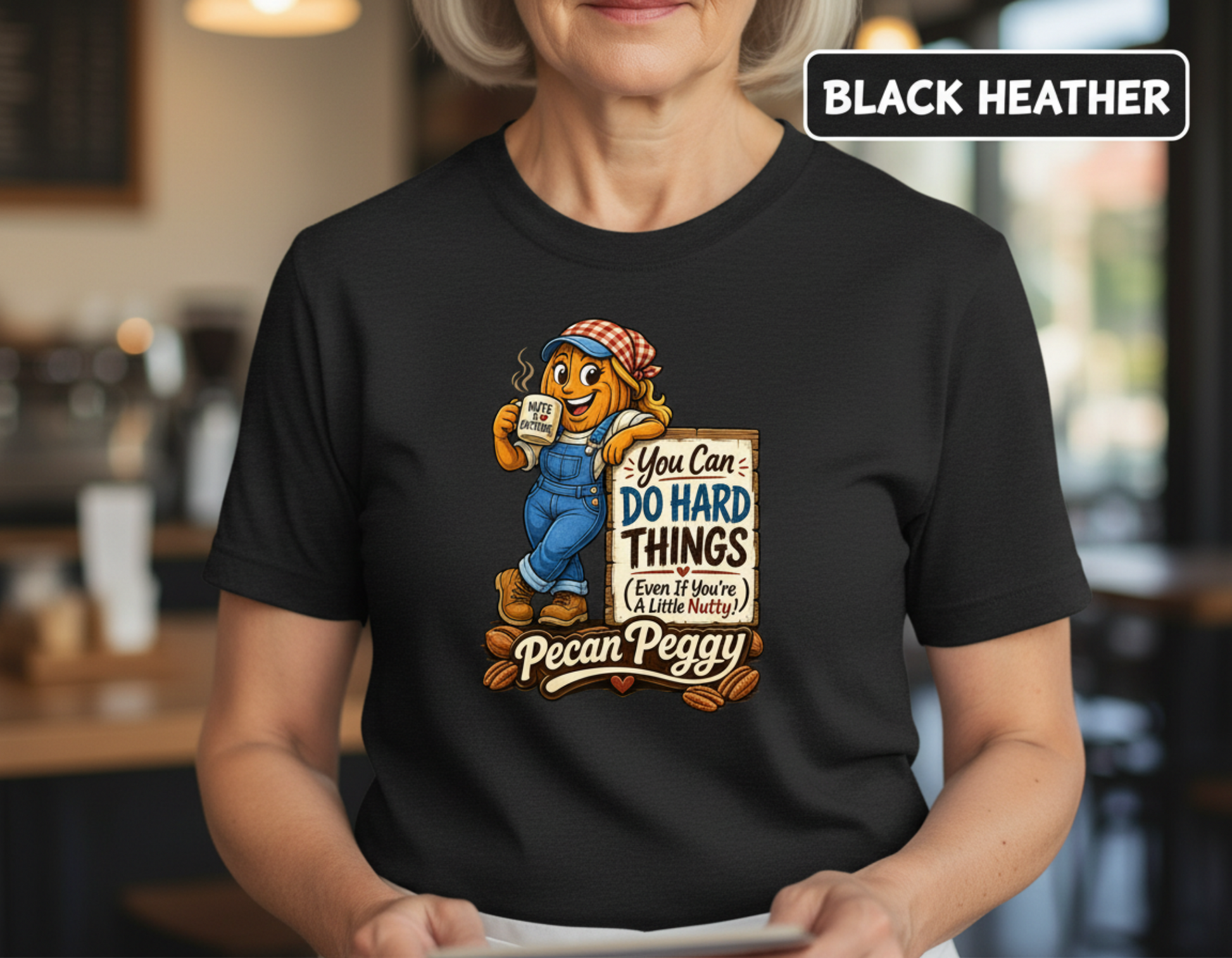 A woman wearing a black graphic t-shirt featuring the character Pecan Peggy with the text 'You Can: DO HARD THINGS Even If You're A Little Lazy!' and 'Pecan Peggy' written below. The image has a 'BLACK HEATHER' label overlay in the top right corner, and the background is a blurred indoor cafe environment.