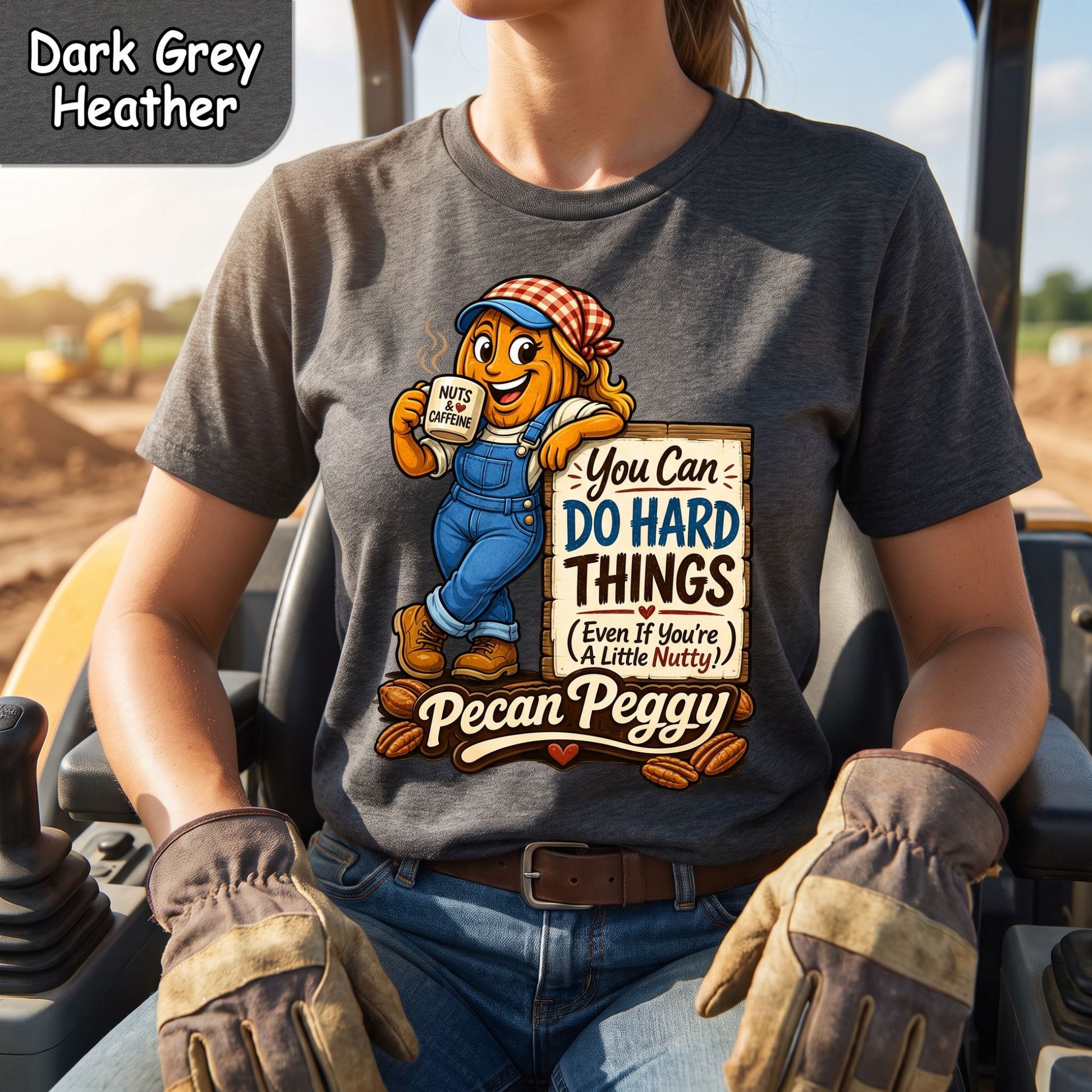 A person wearing a dark grey heather graphic t-shirt with a cartoon pecan character named Pecan Peggy holding a sign that reads 'You Can DO HARD THINGS (Even If You're A Little Nutty!)' and 'Pecan Peggy'. The person is sitting in a construction vehicle with work gloves, with a color swatch label in the top left corner reading 'Dark Grey Heather'.