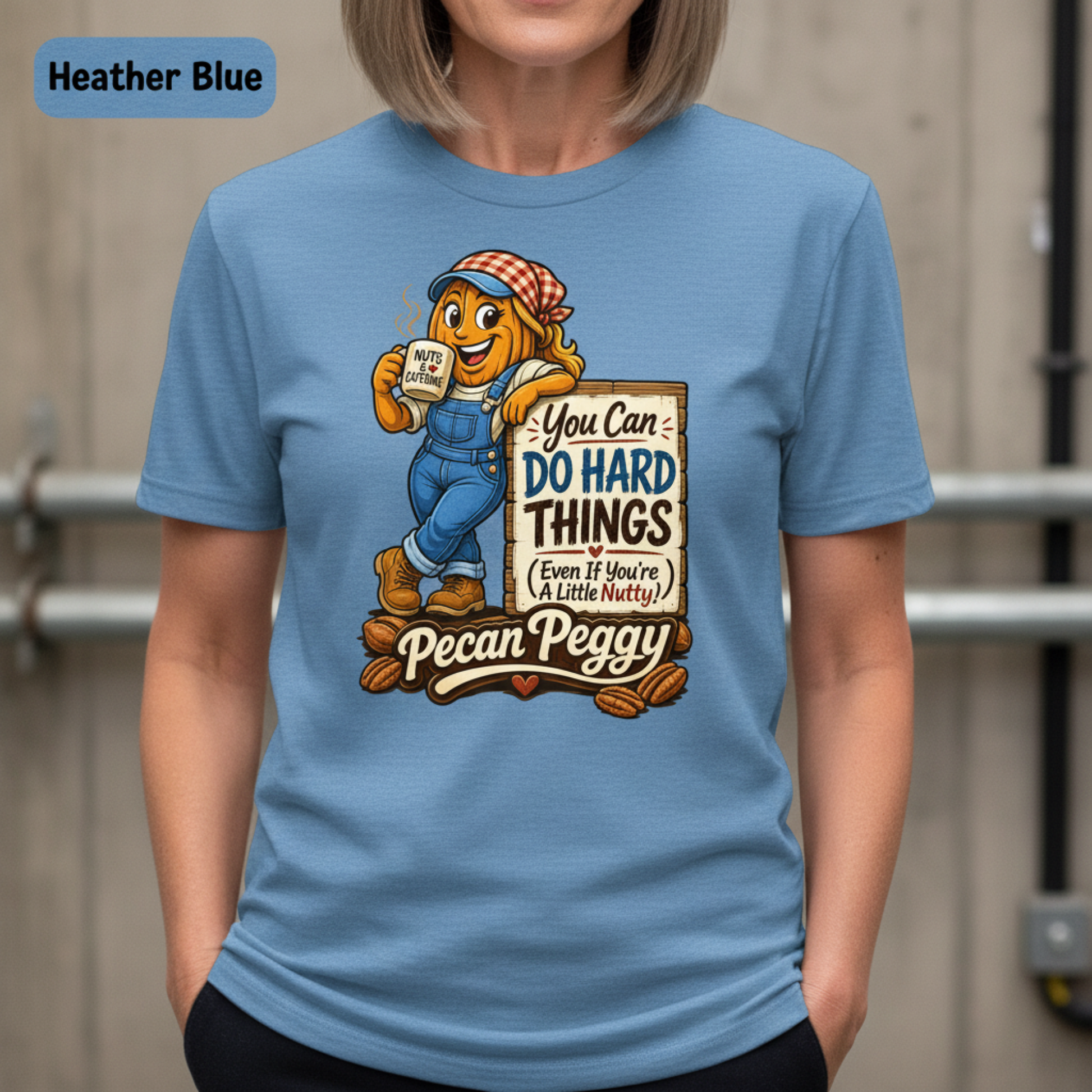 A person wearing a light blue graphic T-shirt with a cartoon pecan character and the text 'You Can DO HARD THINGS (Even If You're A Little Nutty!)' and 'Pecan Peggy.' A color label in the top left reads Heather Blue.