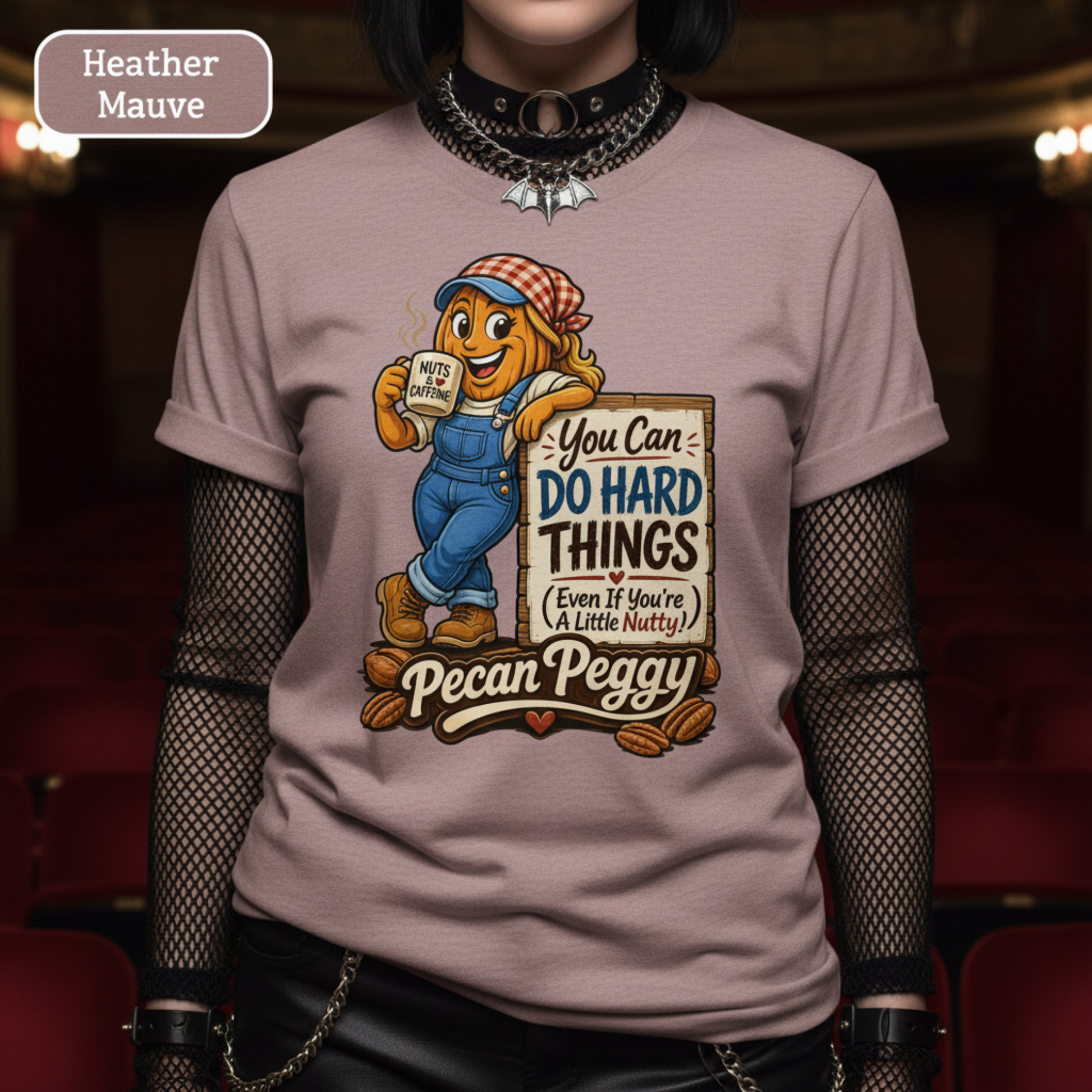 A person wearing a heather mauve graphic t-shirt featuring a cartoon pecan character holding a mug with the text 'You Can DO HARD THINGS (Even If You're A Little Nutsy)' and 'Pecan Peggy' below. The background is a dimly lit, blurred theater or auditorium setting.