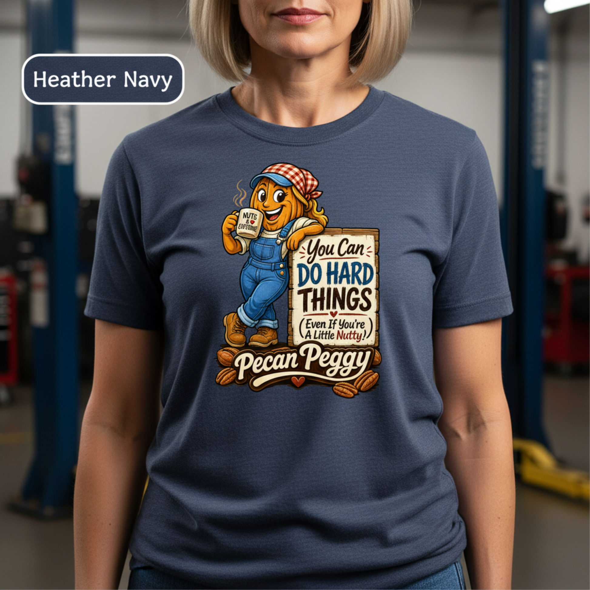 A woman wearing a heather navy graphic t-shirt with a cartoon pecan character and the text 'You Can DO HARD THINGS Even If You're A Little Nasty! Pecan Peggy'. The background is a blurred garage setting.