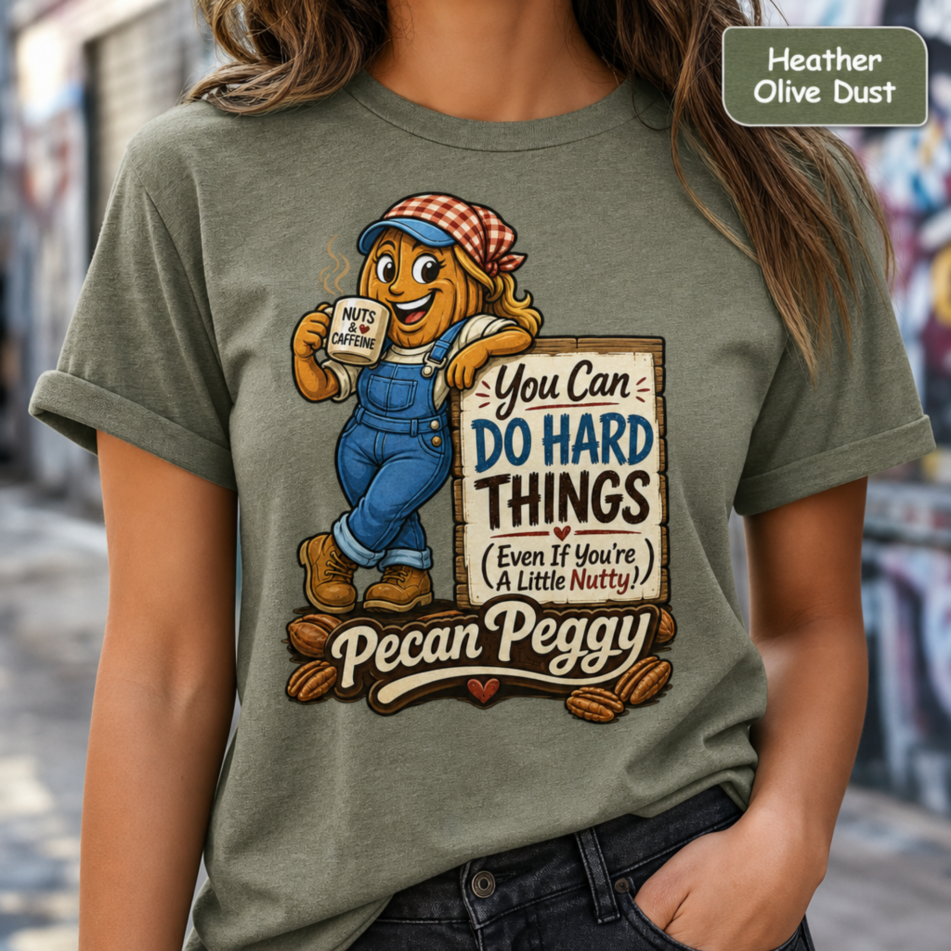 A woman wearing a heather olive dust graphic t-shirt featuring a cartoon pecan character named Pecan Peggy holding a sign that reads 'You Can DO HARD THINGS Even If You're A Little Nutty!' and holding nuts. The background is a blurred outdoor street scene.