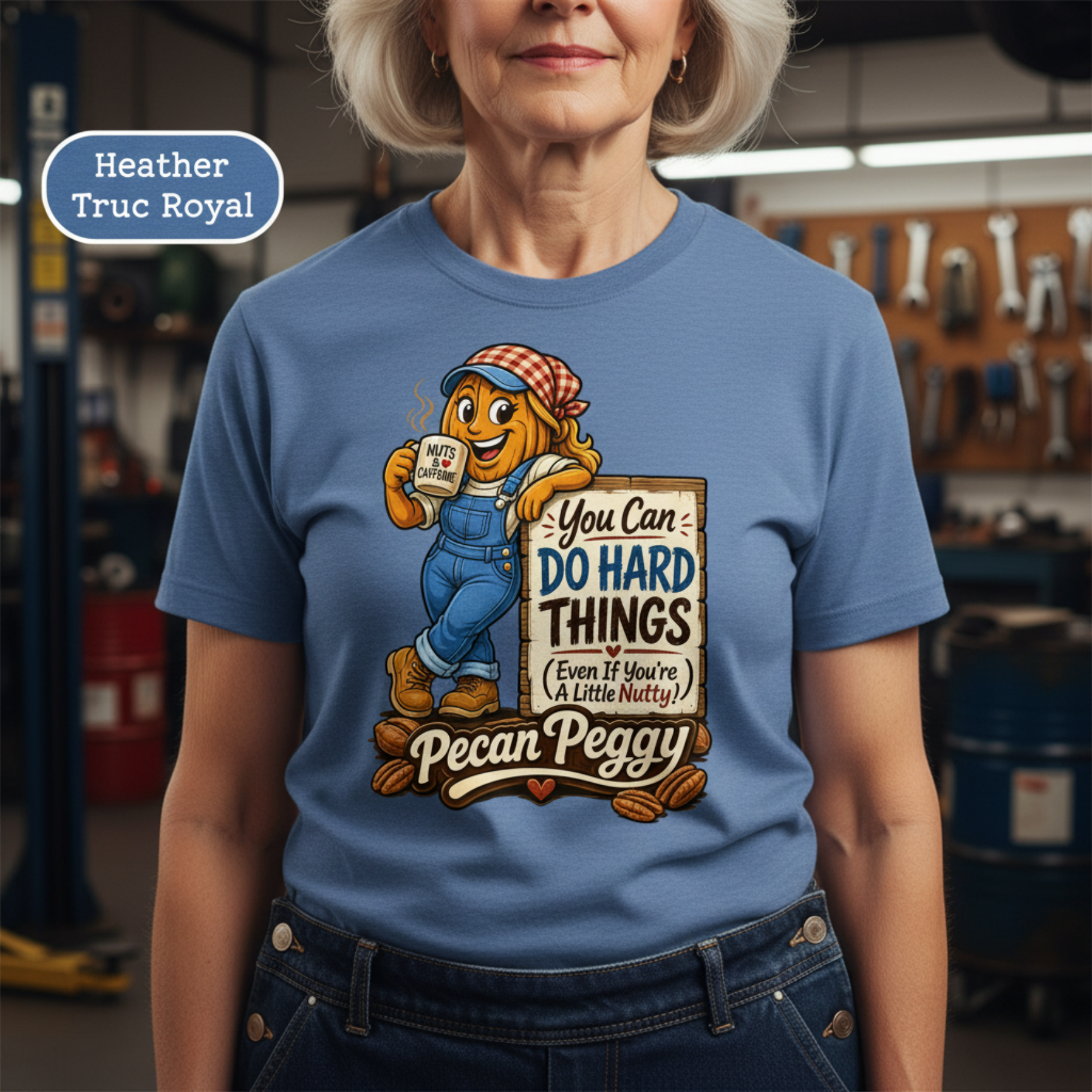 A woman wearing a blue graphic T-shirt with a cartoon pecan character and the text 'Heather Truc Royal' and 'Pecan Peggy'. The background is a blurred garage or workshop setting.
