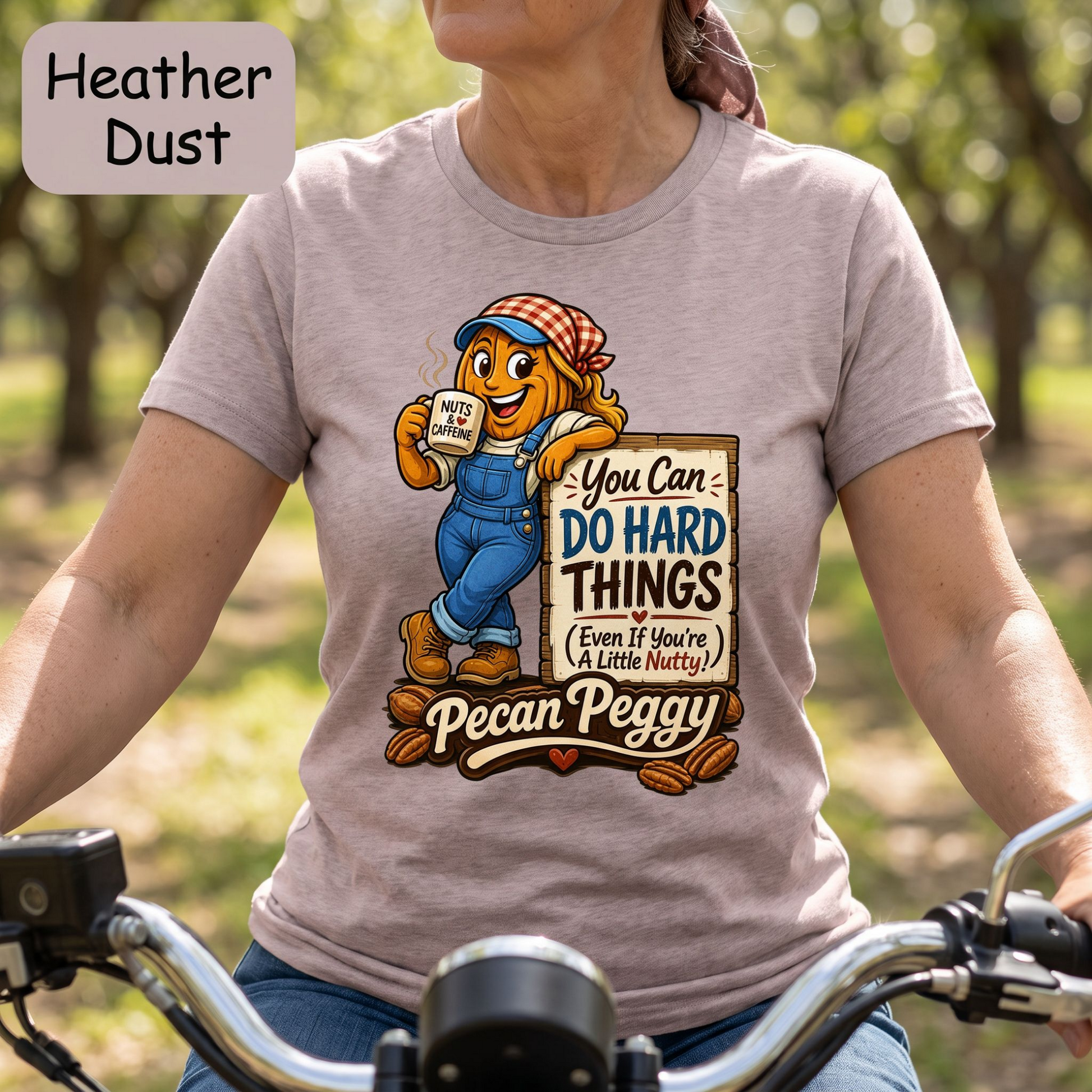 A woman wearing a light heather dusty pink graphic t-shirt while riding a bicycle. The shirt features a cartoon pecan character in blue overalls holding a coffee mug with the text 'My Momma's Coffee is the Best in the World!'.