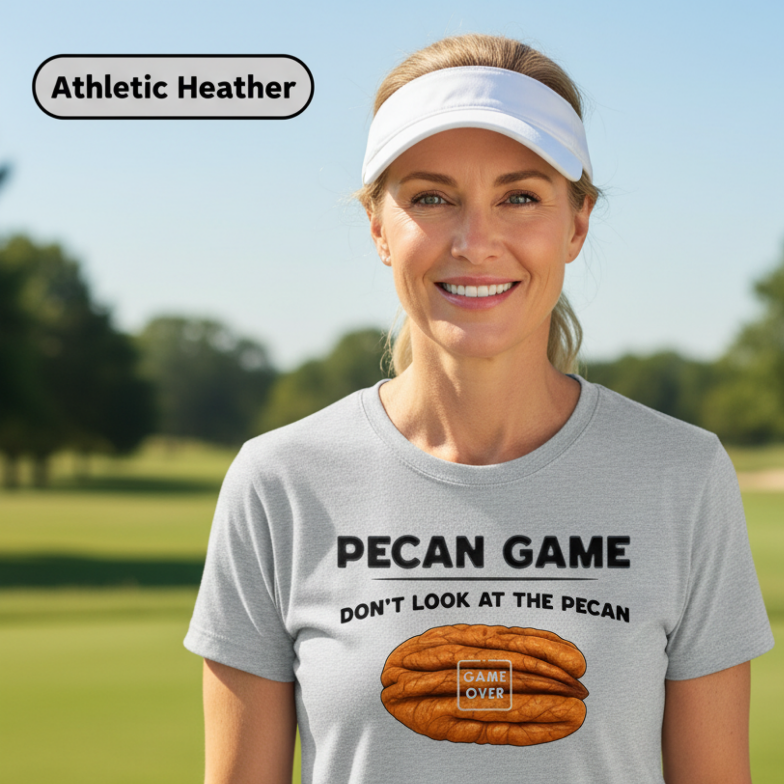 A woman wearing a light heather gray athletic t-shirt with a 'Pecan Game' graphic, standing on a blurred golf course background. The text 'Athletic Heather' is displayed in a label at the top left.