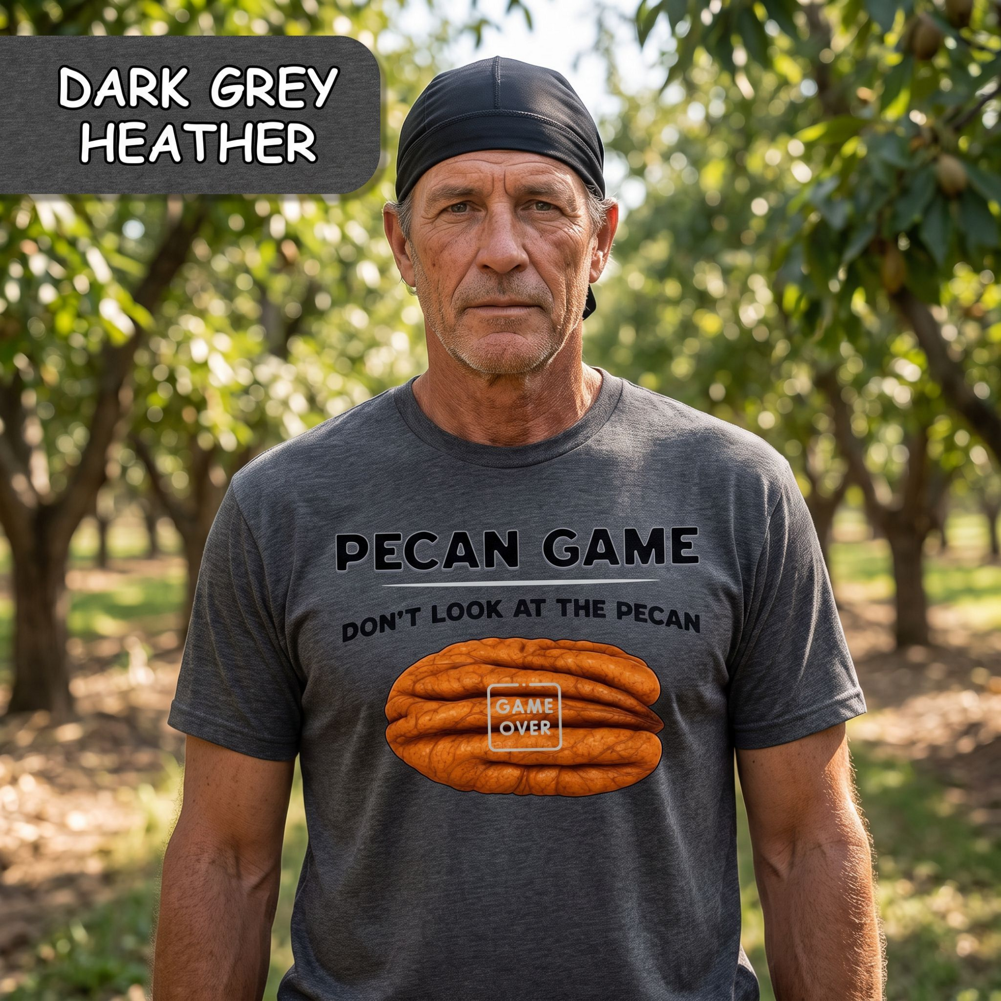 A man standing in a tree-lined outdoor setting wearing a dark grey heather graphic t-shirt with a 'Pecan Game' print. A dark grey label in the top left corner reads 'DARK GREY HEATHER'.