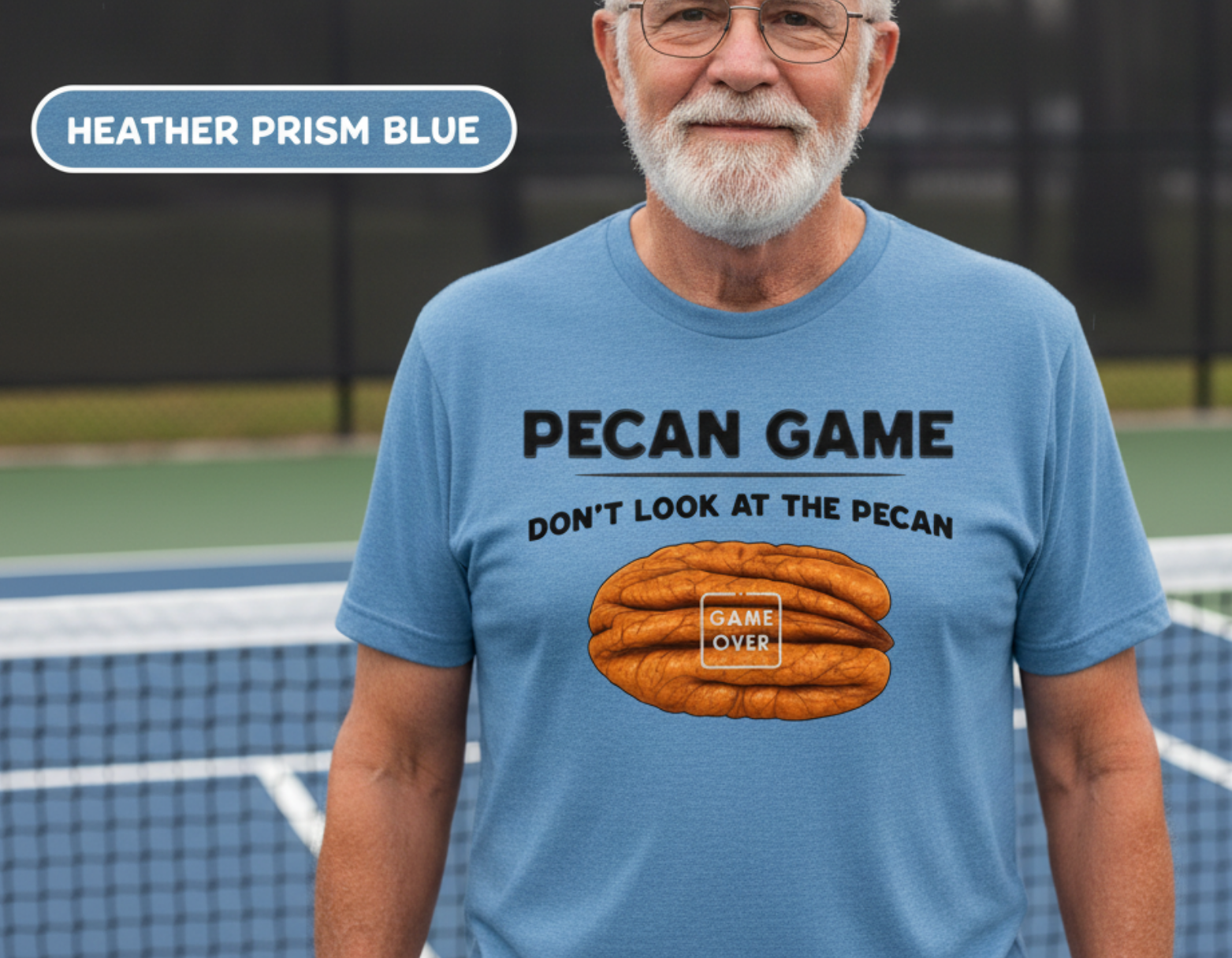 An older man with a white beard stands on a tennis court wearing a heather prism blue t-shirt with a 'Pecan Game' graphic. A tennis ball and racket are visible in the background, and a label in the top left reads 'HEATHER PRISM BLUE'.