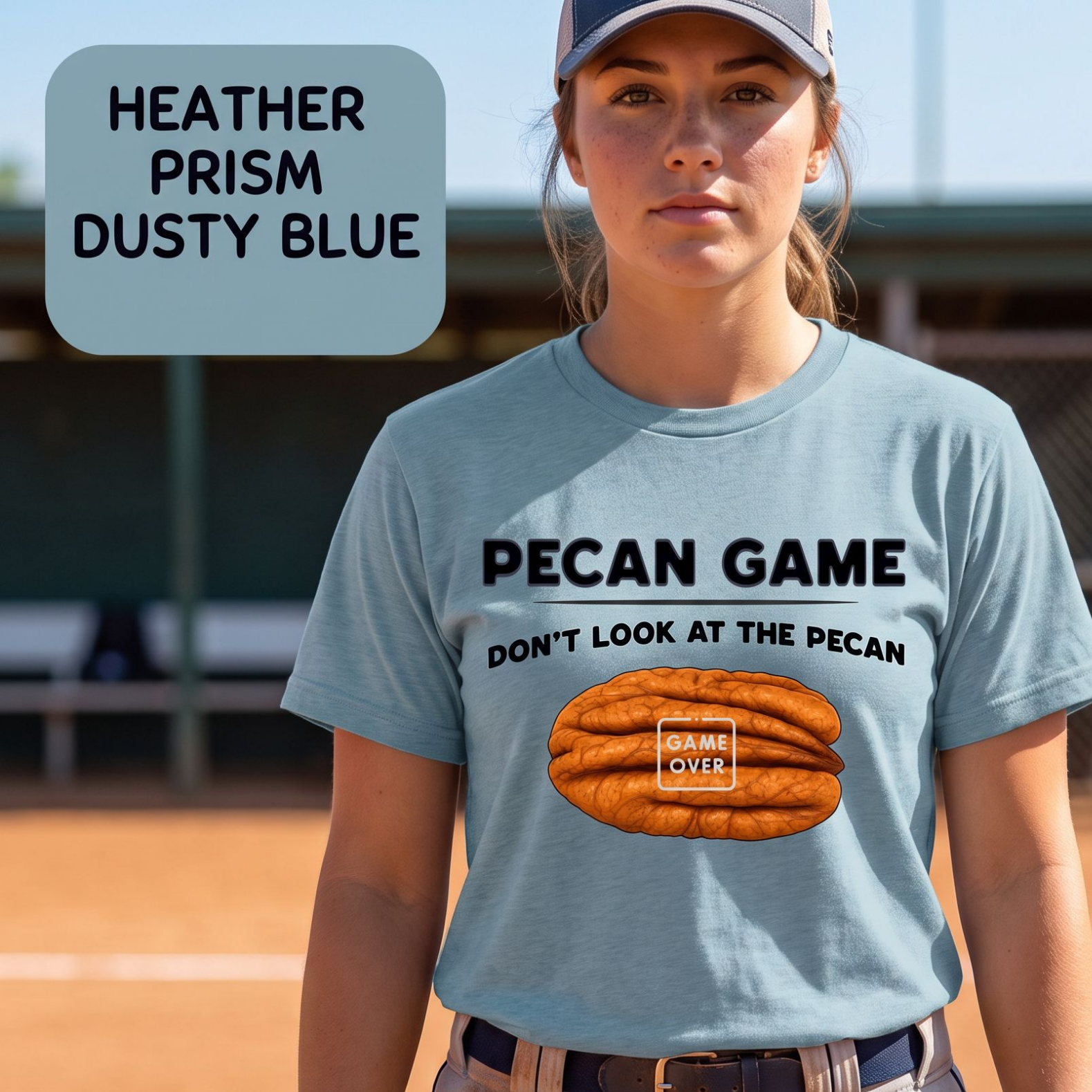 A woman wearing a heather prism dusty blue graphic t-shirt on a baseball field. The shirt features a black text graphic reading 'PECAN GAME DON'T LOOK AT THE PECAN' above an illustration of a pecan with 'GAME OVER' written on it.