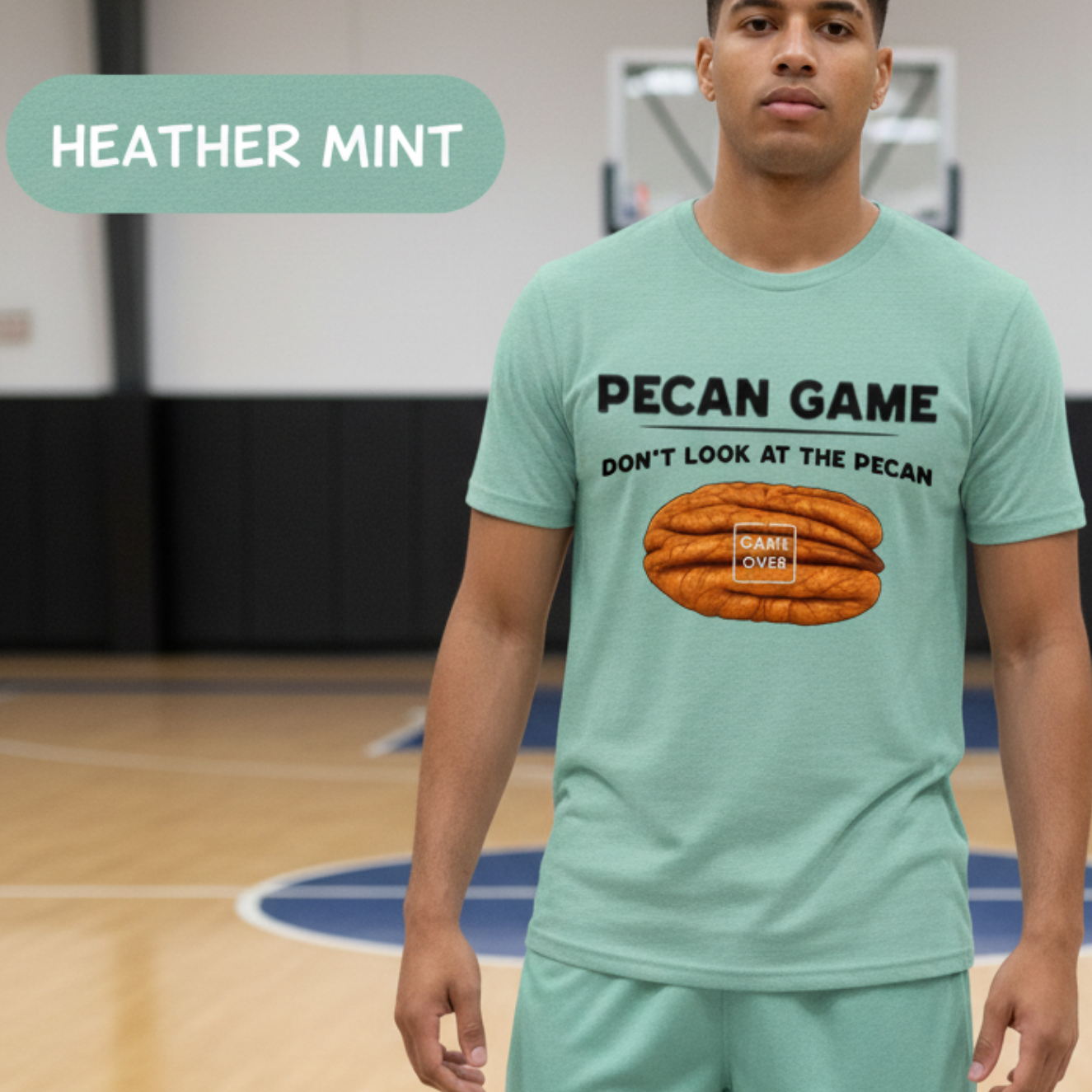 A man standing on an indoor basketball court wearing a heather mint green athletic t-shirt and matching shorts. The t-shirt features a graphic of a pecan with the text 'PECAN GAME DON'T LOOK AT THE PECAN'.