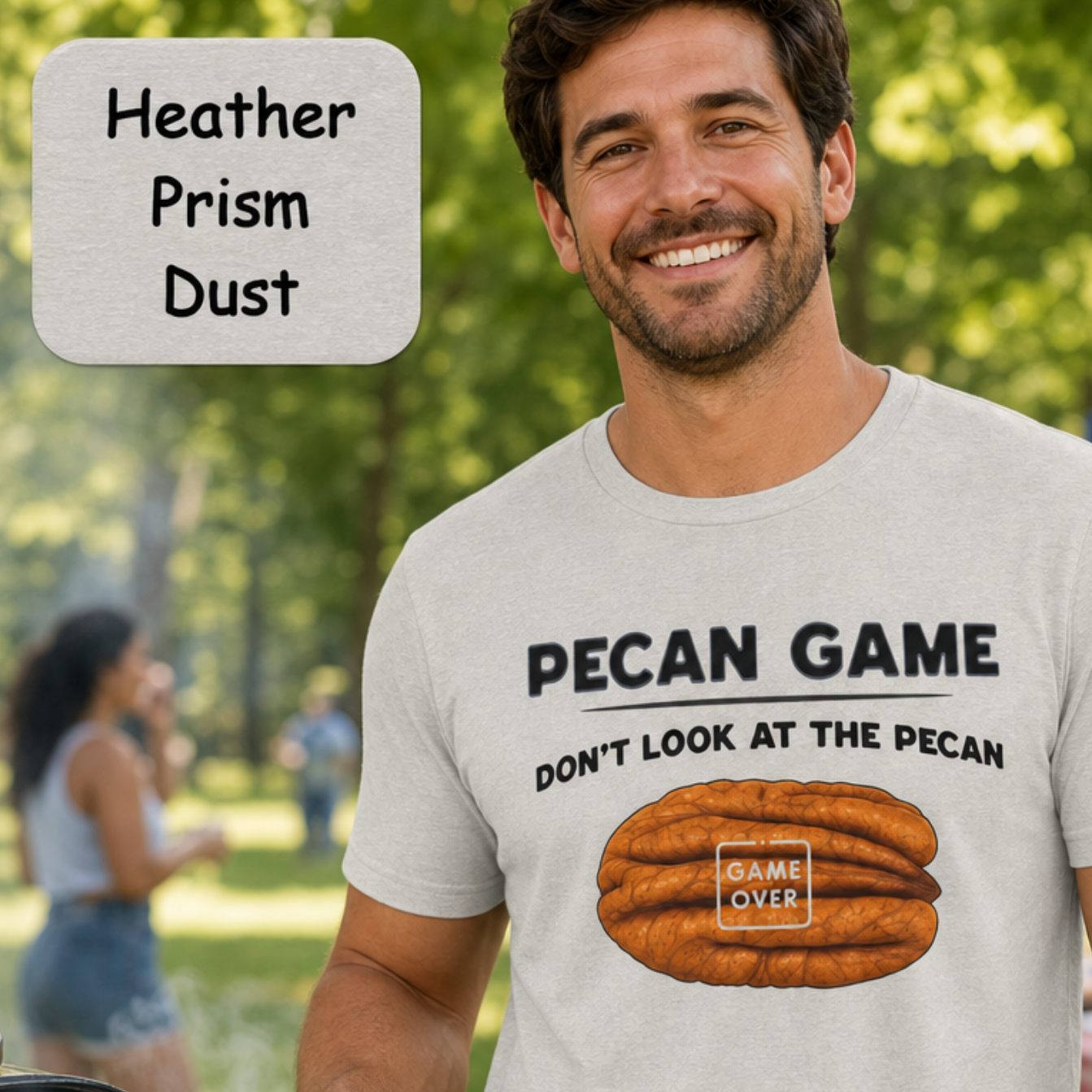 A man at an outdoor barbecue wearing a light heather gray t-shirt with a humorous graphic that reads 'PECAN GAME DON'T LOOK AT THE PECAN' above an illustration of a pecan nut. A text overlay in the top left corner reads 'Heather Prism Dust'.