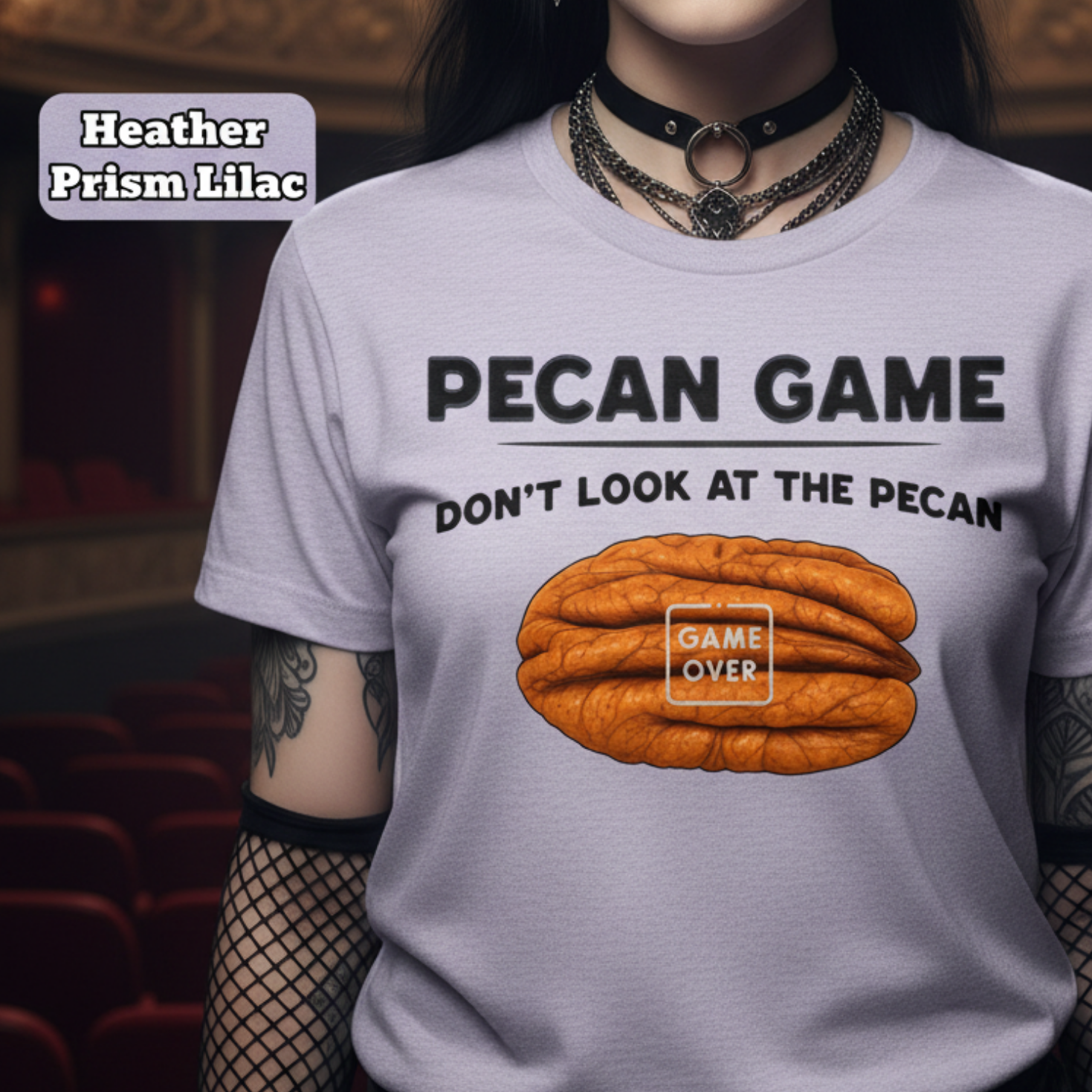A model wearing a light lavender graphic t-shirt with a 'Pecan Game' design, set against a dark, gothic-themed theater background. The shirt features a graphic of an orange pecan with the words 'GAME OVER' and 'DON'T LOOK AT THE PECAN'.