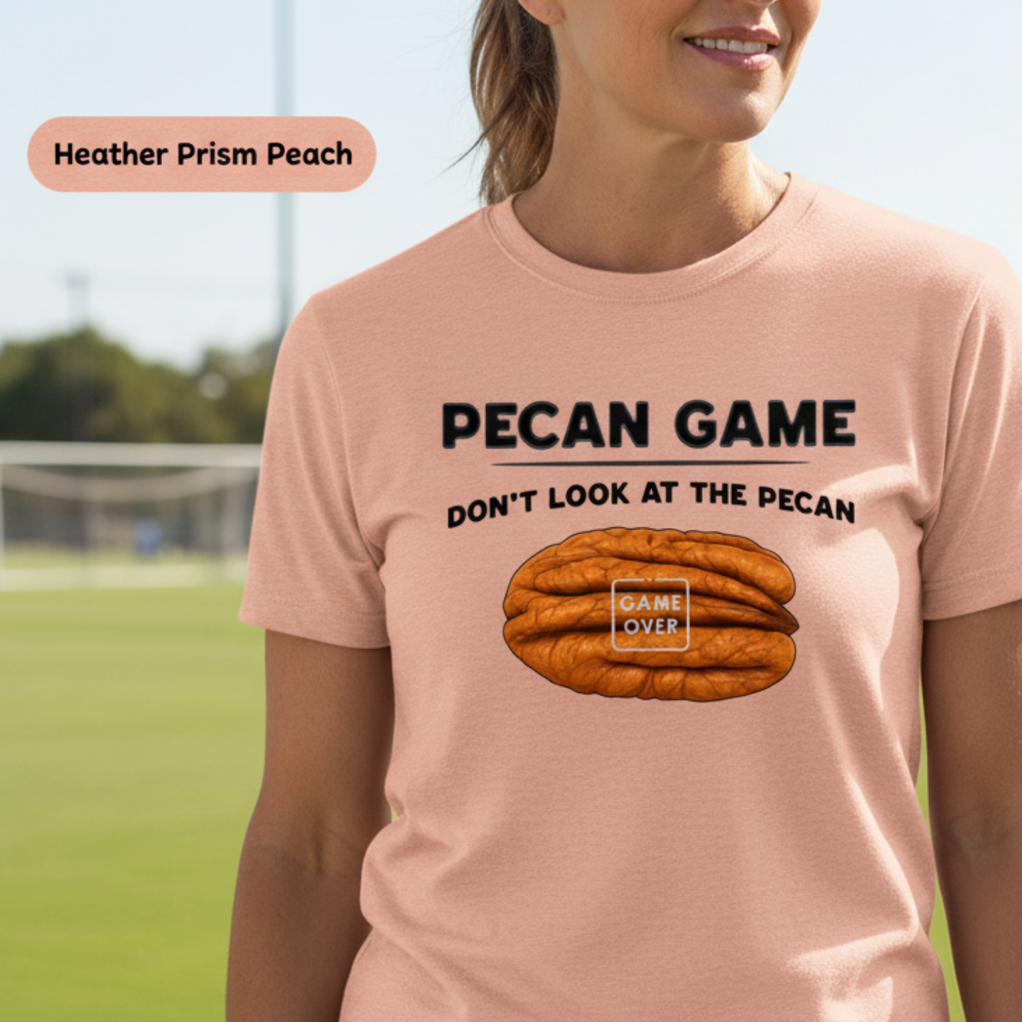 A woman wearing a light peach graphic t-shirt with a 'Pecan Game' design, standing on a green soccer field. The t-shirt features a graphic of a pecan nut with the words 'DON'T LOOK AT THE PECAN' and 'GAME OVER' written on it.