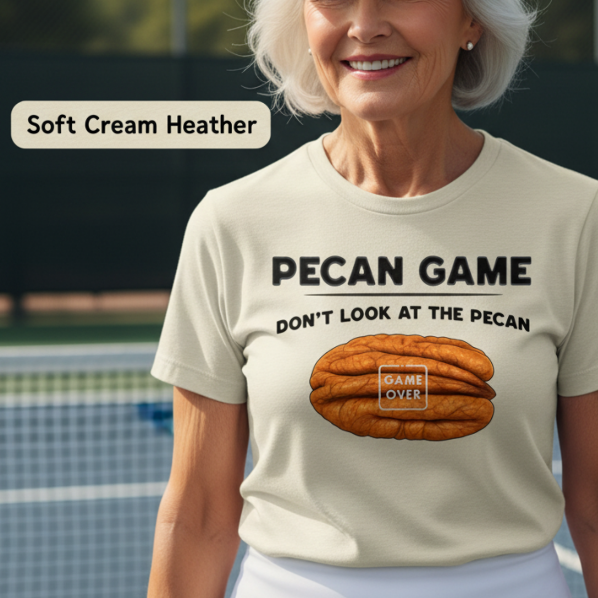 A smiling older woman on a tennis court wearing a cream heather graphic T-shirt with the text 'PECAN GAME DON'T LOOK AT THE PECAN' and a pecan illustration. A small label in the upper left reads 'Soft Cream Heather'.