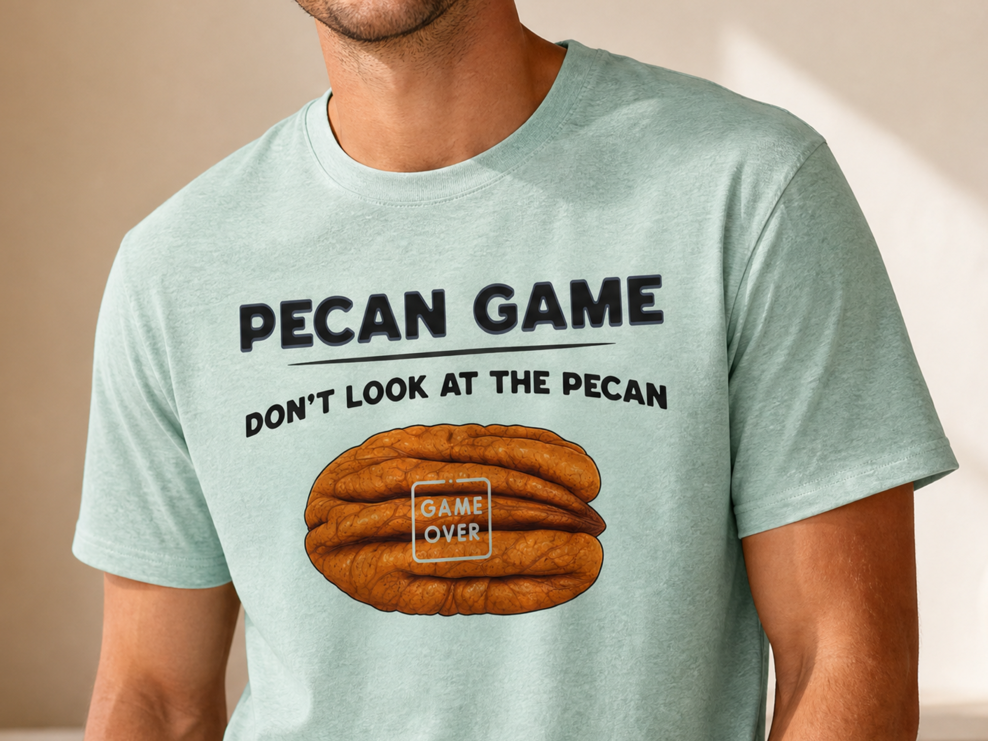 A smiling man wearing a light mint green graphic t-shirt with a brown pecan illustration and the text 'PECAN GAME DON'T LOOK AT THE PECAN' on the front, set in a warm, sunlit room.