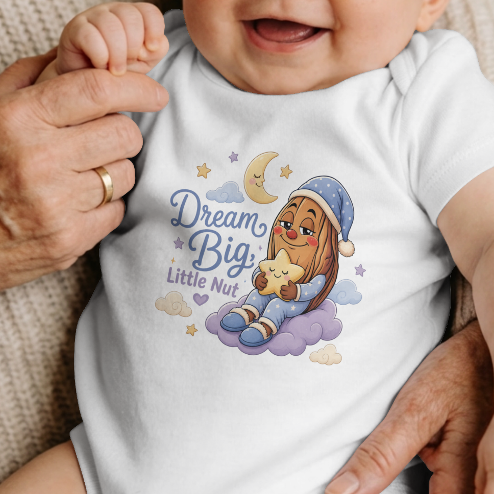 A smiling baby with red hair sits on an adult's lap, wearing a white short-sleeve bodysuit with a colorful cartoon illustration and the text 'Dream Big Little My'.