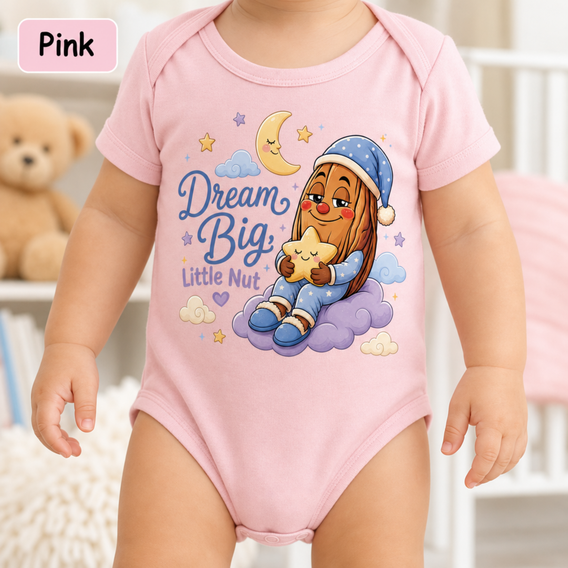 A baby wearing a pink short-sleeve bodysuit with a cartoon graphic of a sleeping nut under a crescent moon and the text 'Dream Big Little Nut'. A pink label in the top left corner reads 'Pink'.