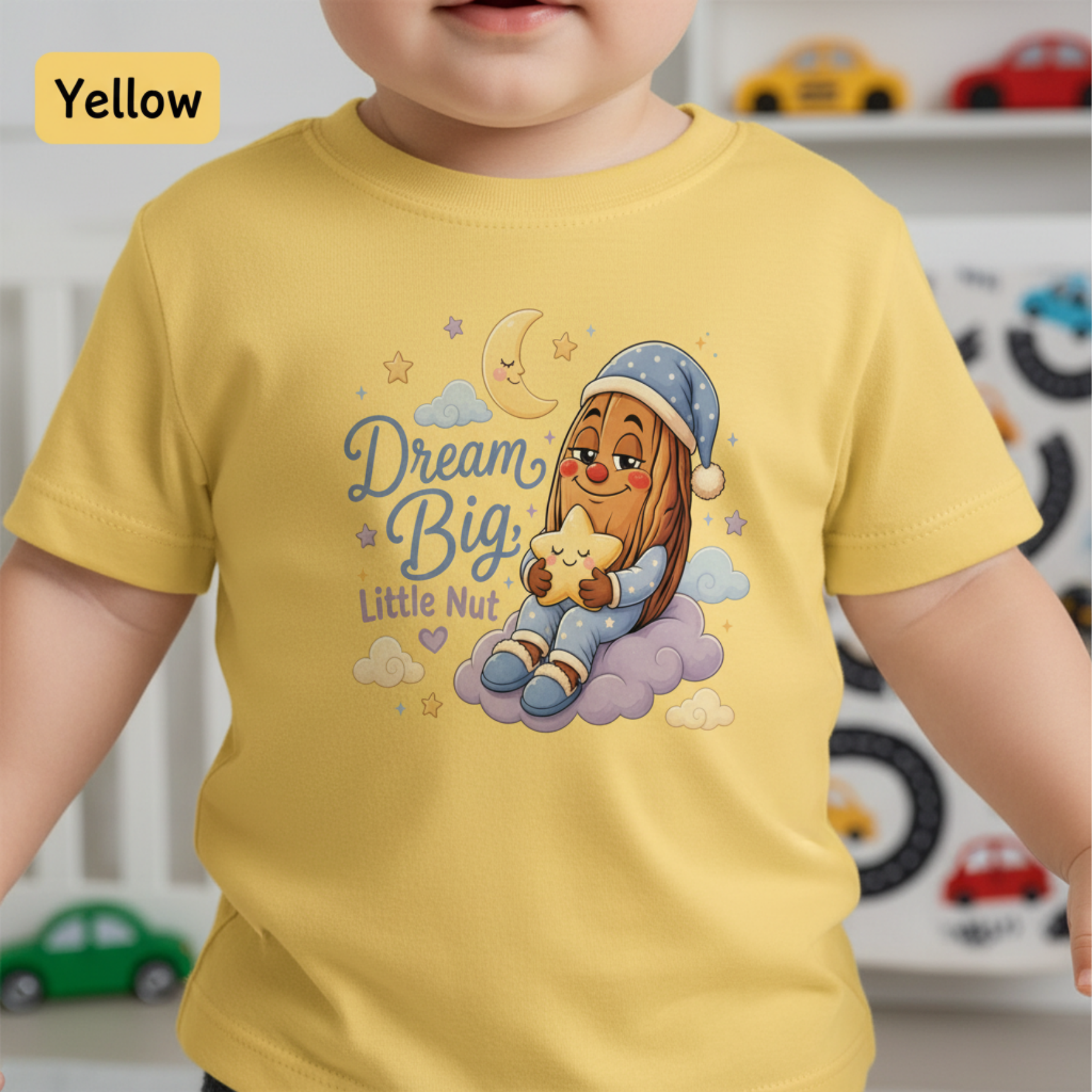 A toddler wearing a pale yellow short-sleeve t-shirt with a colorful cartoon graphic of a girl in a blue nightcap and pajamas holding a star on a cloud, next to the text 'Dream Big, Little Nut'. A 'Yellow' color label is visible in the top left corner.