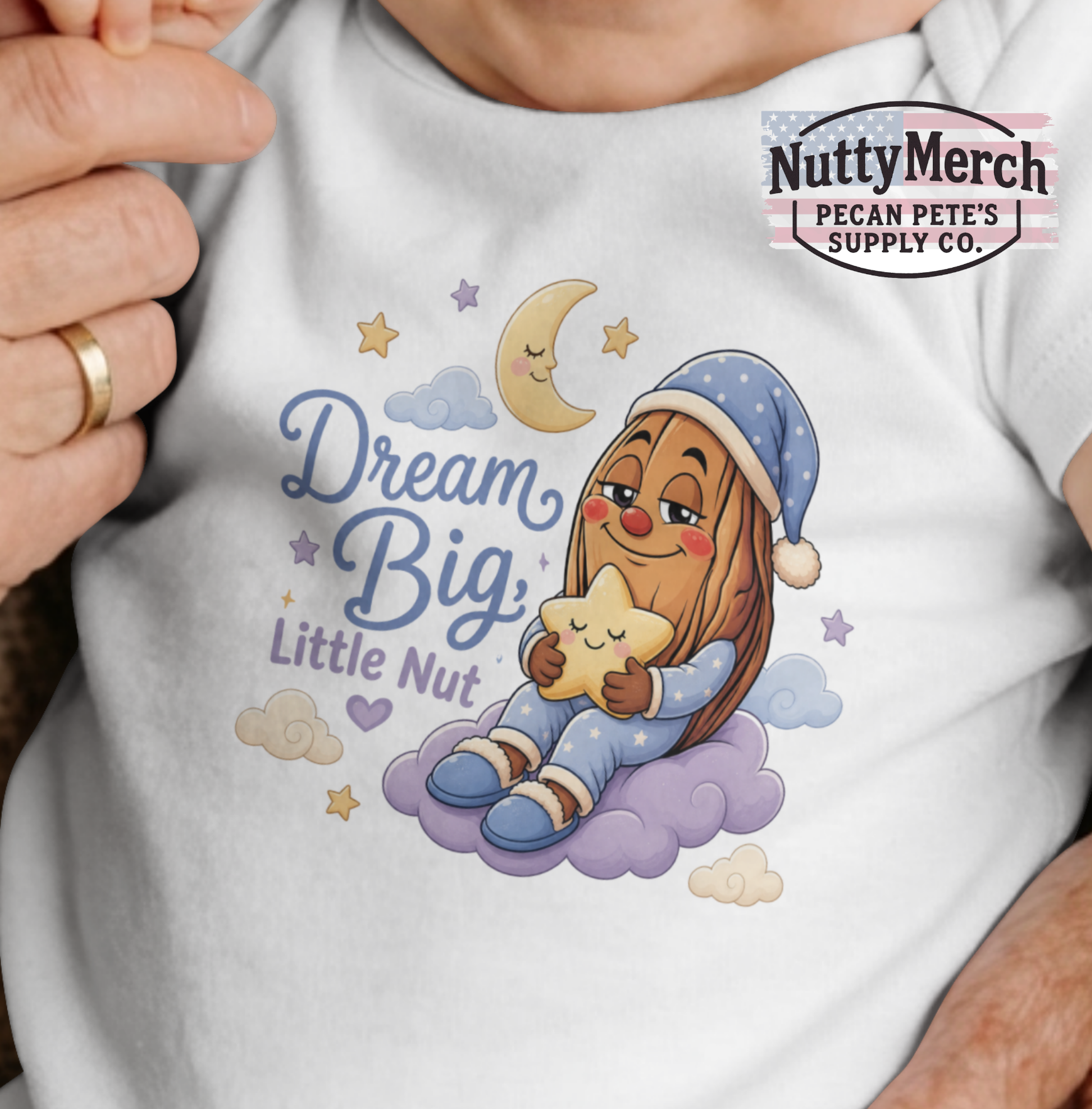 A close-up of a white baby or toddler tank top worn by a person, featuring a colorful cartoon graphic of a sleeping girl in a blue starry nightcap and pajamas hugging a star on a cloud, with the text 'Dream Big, Little Nut'. A 'NuttyMerch Pecan Pete's Supply Co.