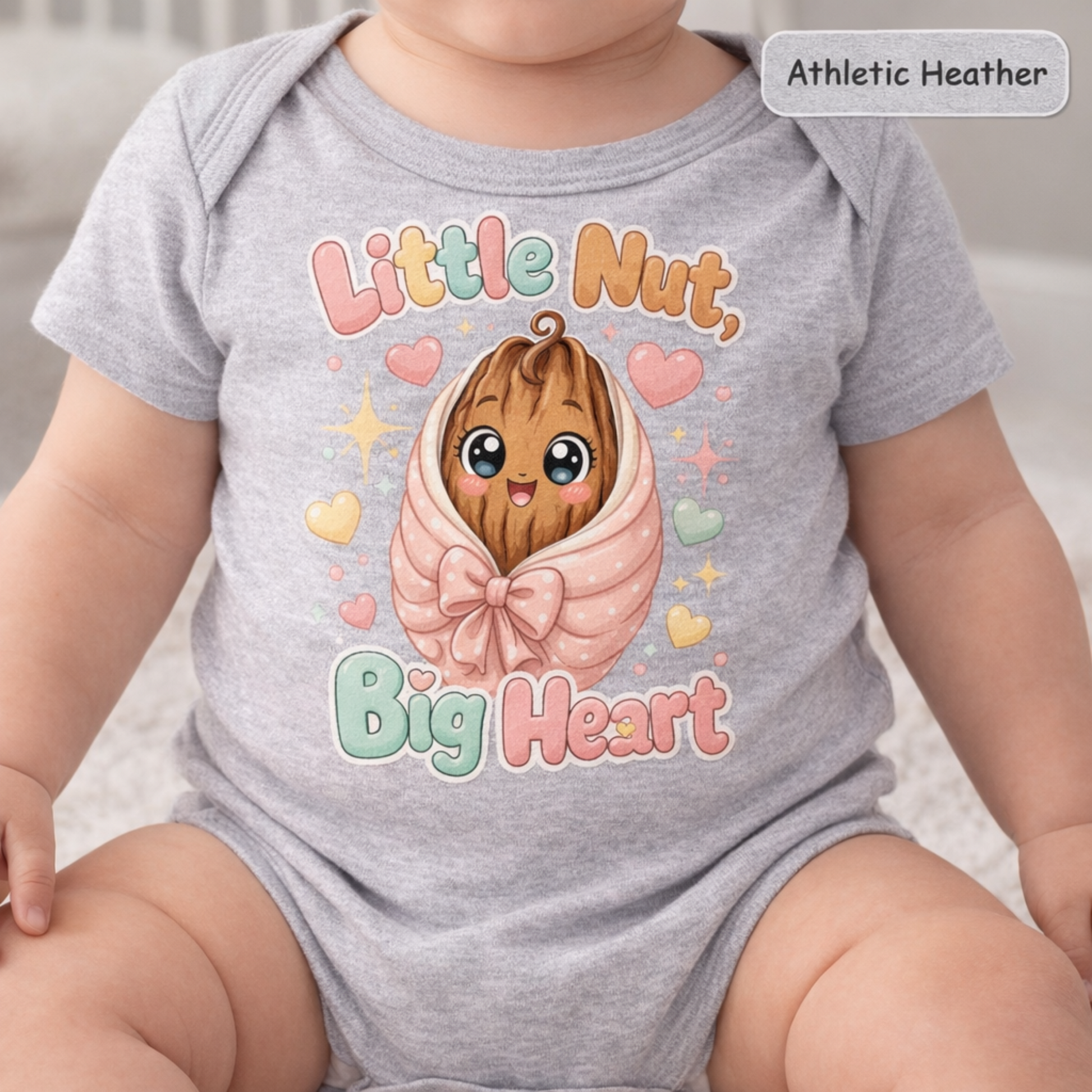 A baby wearing an athletic heather gray short-sleeve bodysuit with a cute graphic of a smiling cartoon nut wrapped in pink fabric, surrounded by pastel hearts and the words 'Little Nut, Big Heart.'.