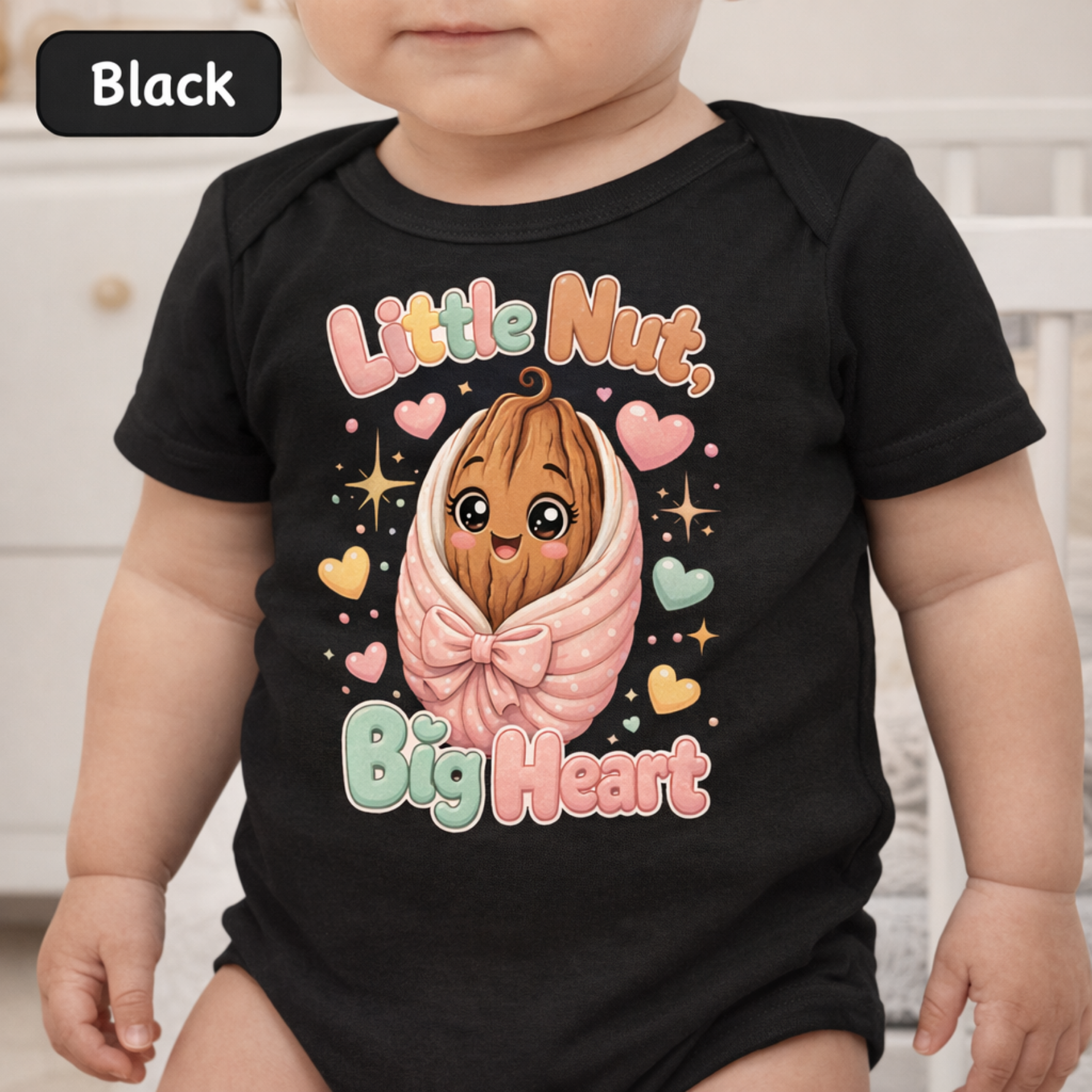 A baby wearing a black short-sleeve bodysuit with a colorful cartoon graphic of a smiling walnut character wrapped in a pink bow, surrounded by hearts and stars, with the text 'Little Nut, Big Heart.' A black label in the top left corner reads 'Black.