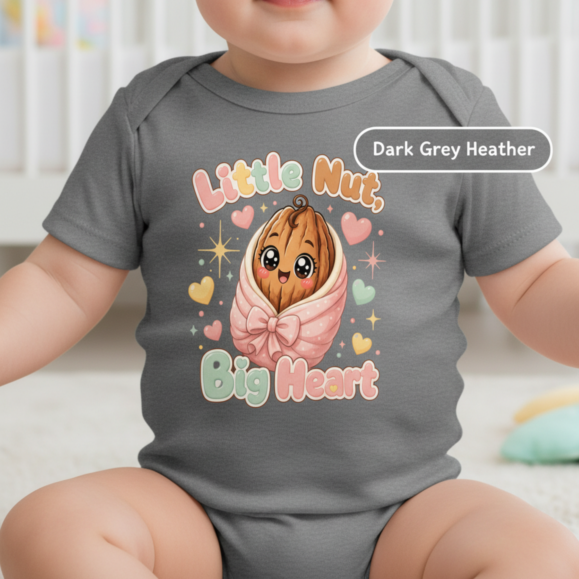 A baby wearing a dark grey heather short-sleeve bodysuit with a cute cartoon graphic of a smiling nut wrapped in pink fabric, surrounded by hearts and the words 'Little Nut Big Heart'. A label overlay reads 'Dark Grey Heather'.