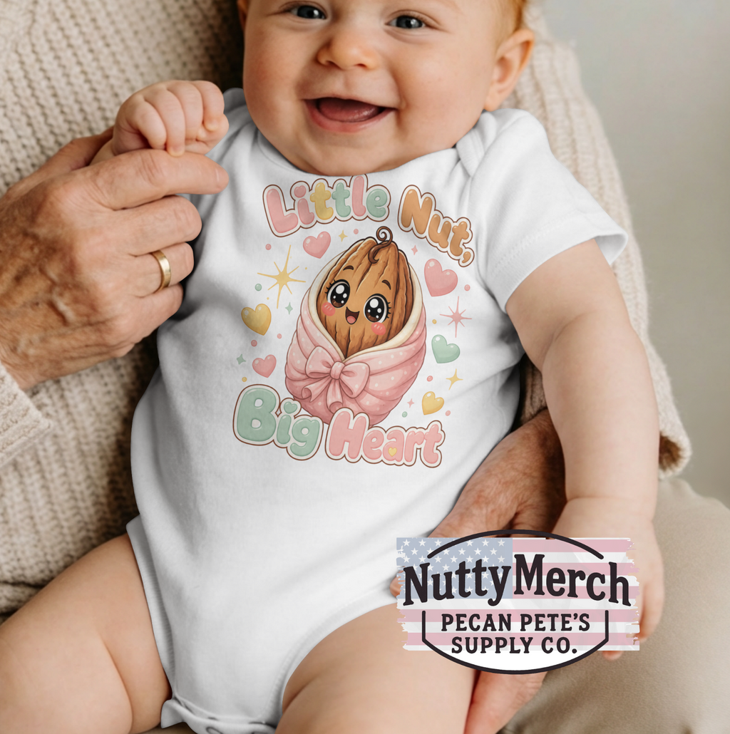 A baby wearing a white short-sleeve bodysuit with a cute cartoon peanut graphic and the text 'Little Nut Big Heart,' held by an adult. A 'NuttyMerch Pecan Pete's Supply Co.