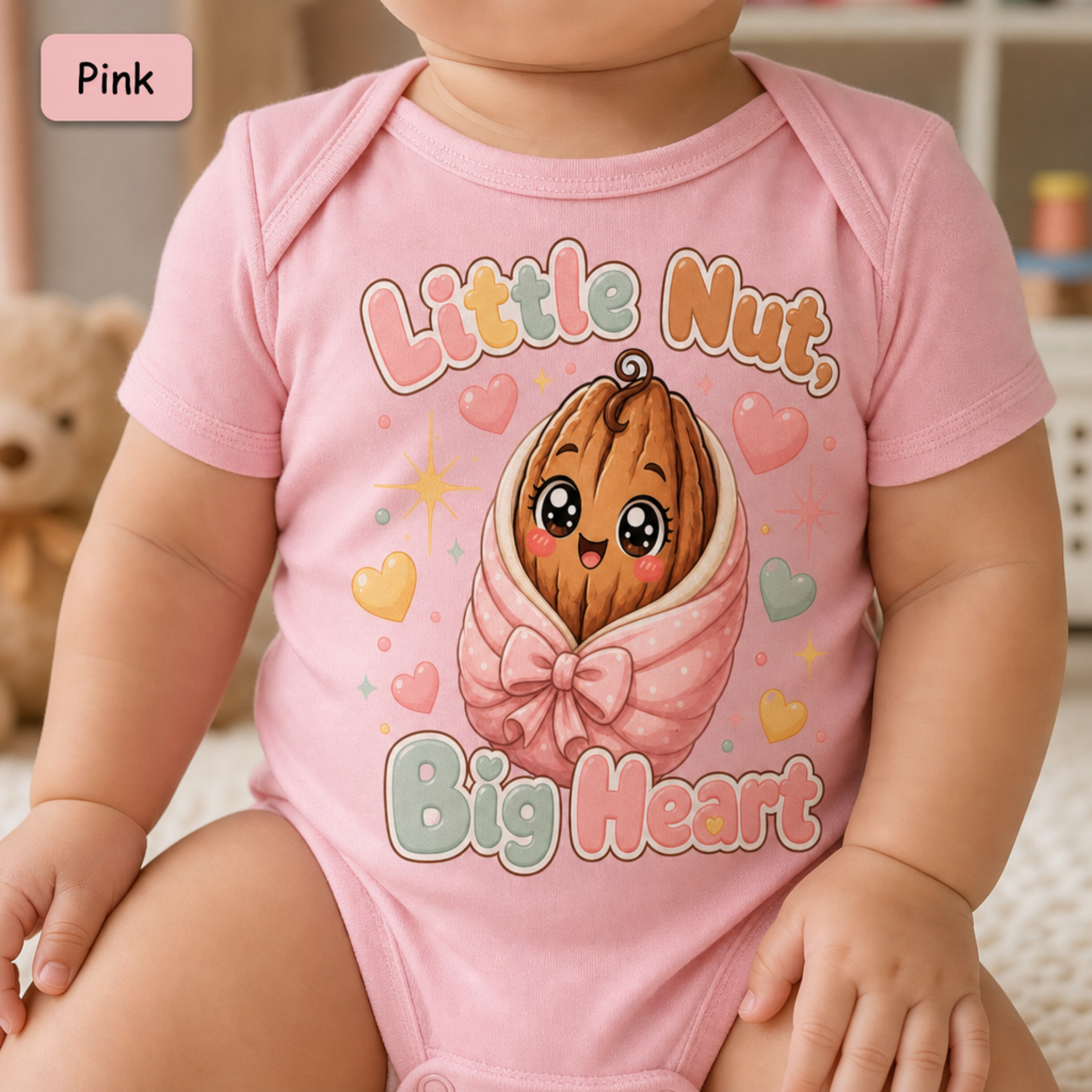 A baby wearing a pink short-sleeve bodysuit with a cute cartoon graphic of a smiling nut wrapped in a pink bow, surrounded by hearts and the text 'Little Nut, Big Heart'. A pink label in the top left corner reads 'Pink'.