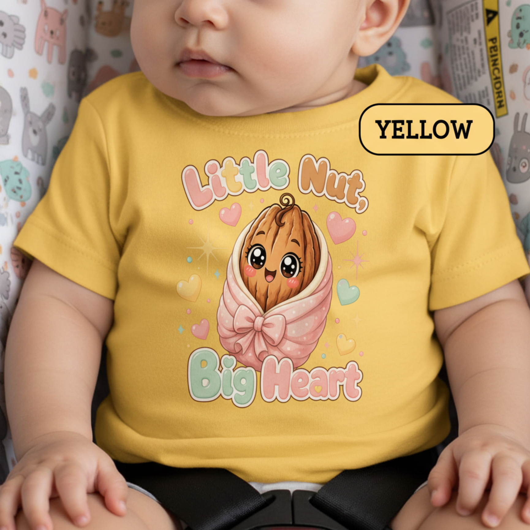 A baby wearing a bright yellow short-sleeve t-shirt with a cute cartoon graphic of a smiling nut wrapped in a pink bow and the text 'Little Nut, Big Heart'. A 'YELLOW' label is visible in the top right corner.