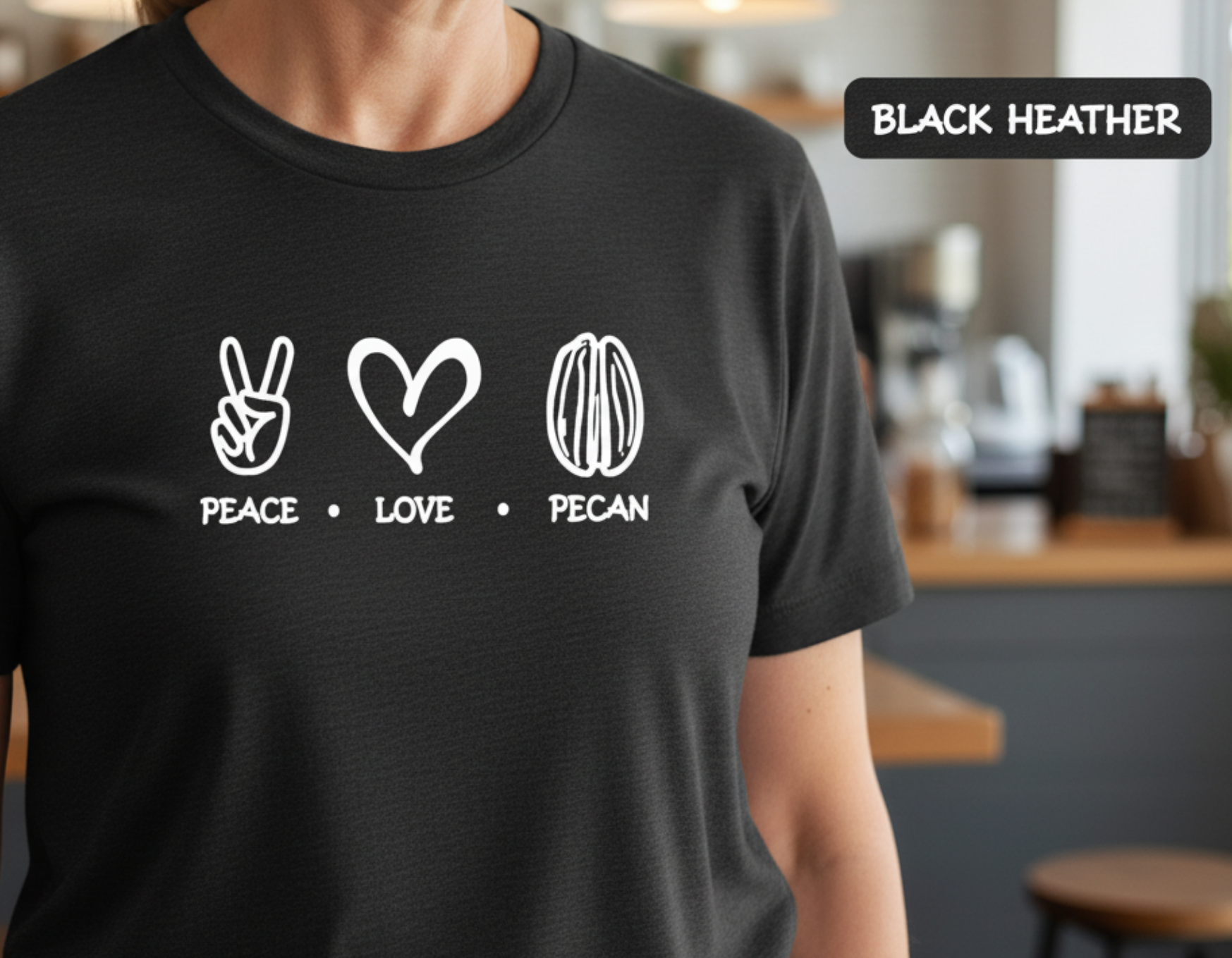 A person wearing a black short-sleeve t-shirt with a white graphic print of peace, love, and pecan symbols with the words 'PEACE • LOVE • PECAN' underneath. A black label in the top right corner reads 'BLACK HEATHER'.