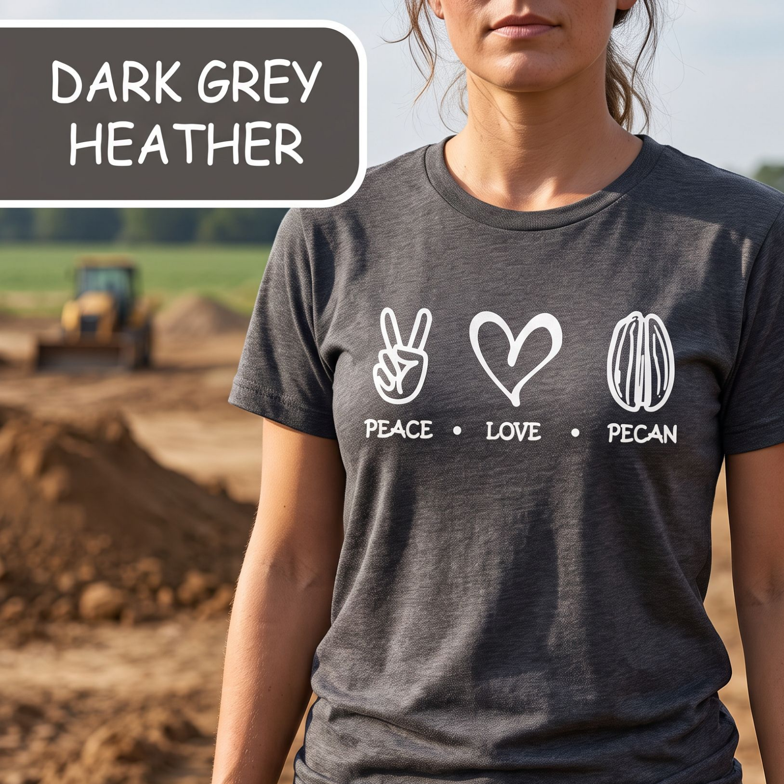A woman wearing a dark grey heather graphic t-shirt with white printed icons and text ('PEACE • LOVE • PECAN') on a dirt construction site. A label in the top-left corner reads 'DARK GREY HEATHER'.
