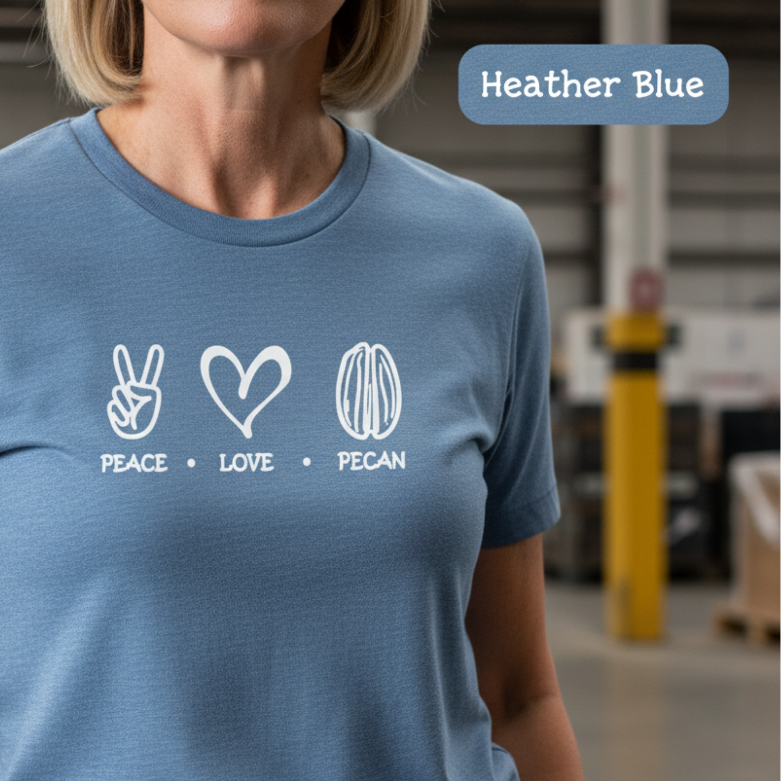 A woman wearing a heather blue short-sleeve t-shirt with a white graphic print that reads 'PEACE • LOVE • PECAN' with peace sign, heart, and pecan nut icons. The background is a blurred warehouse or studio setting.