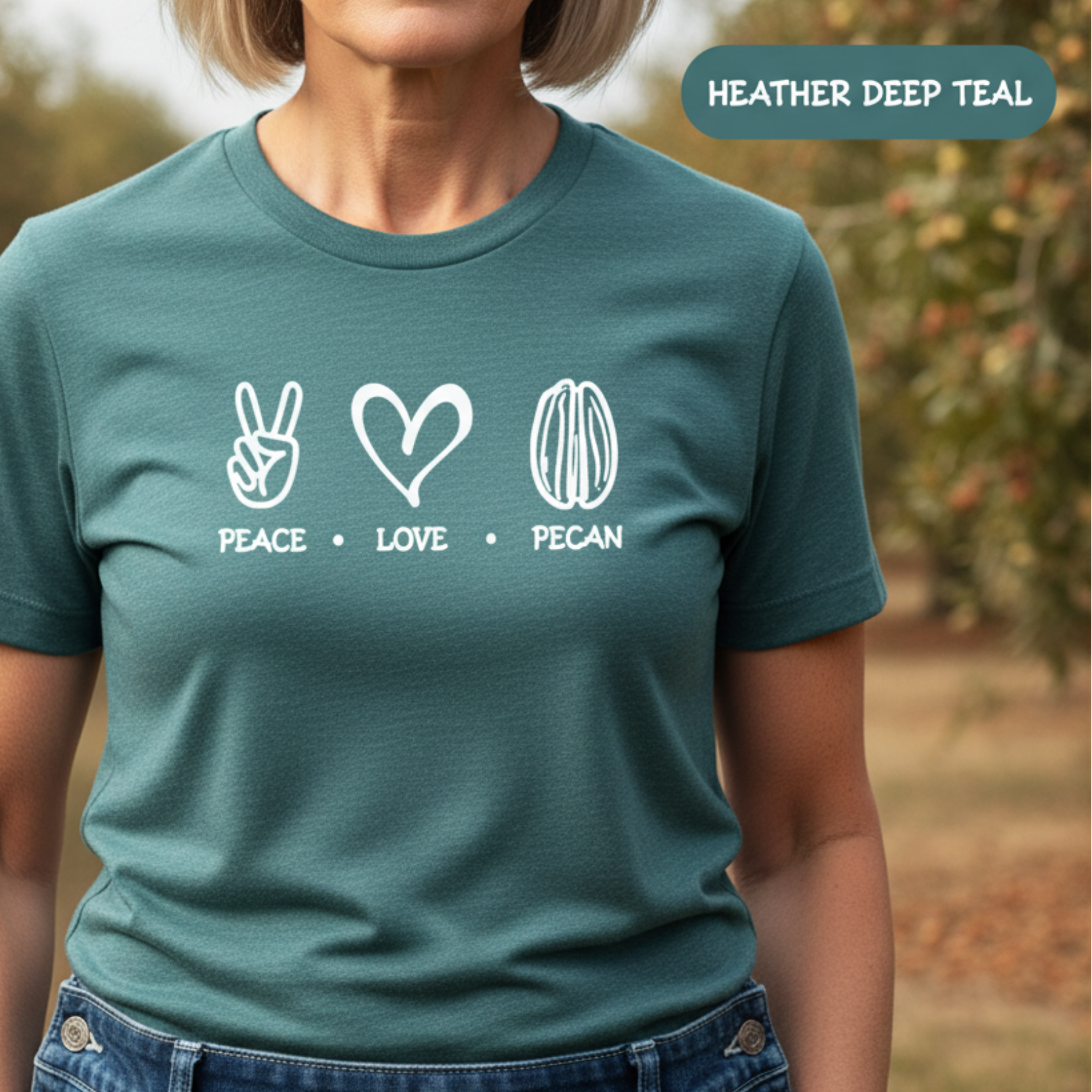 A woman wearing a heather deep teal graphic t-shirt with white line art icons of a peace hand, a heart, and a pecan, with the words 'PEACE', 'LOVE', and 'PECAN' underneath. She is wearing blue jeans and tan work gloves, standing outdoors in a wooded setting.