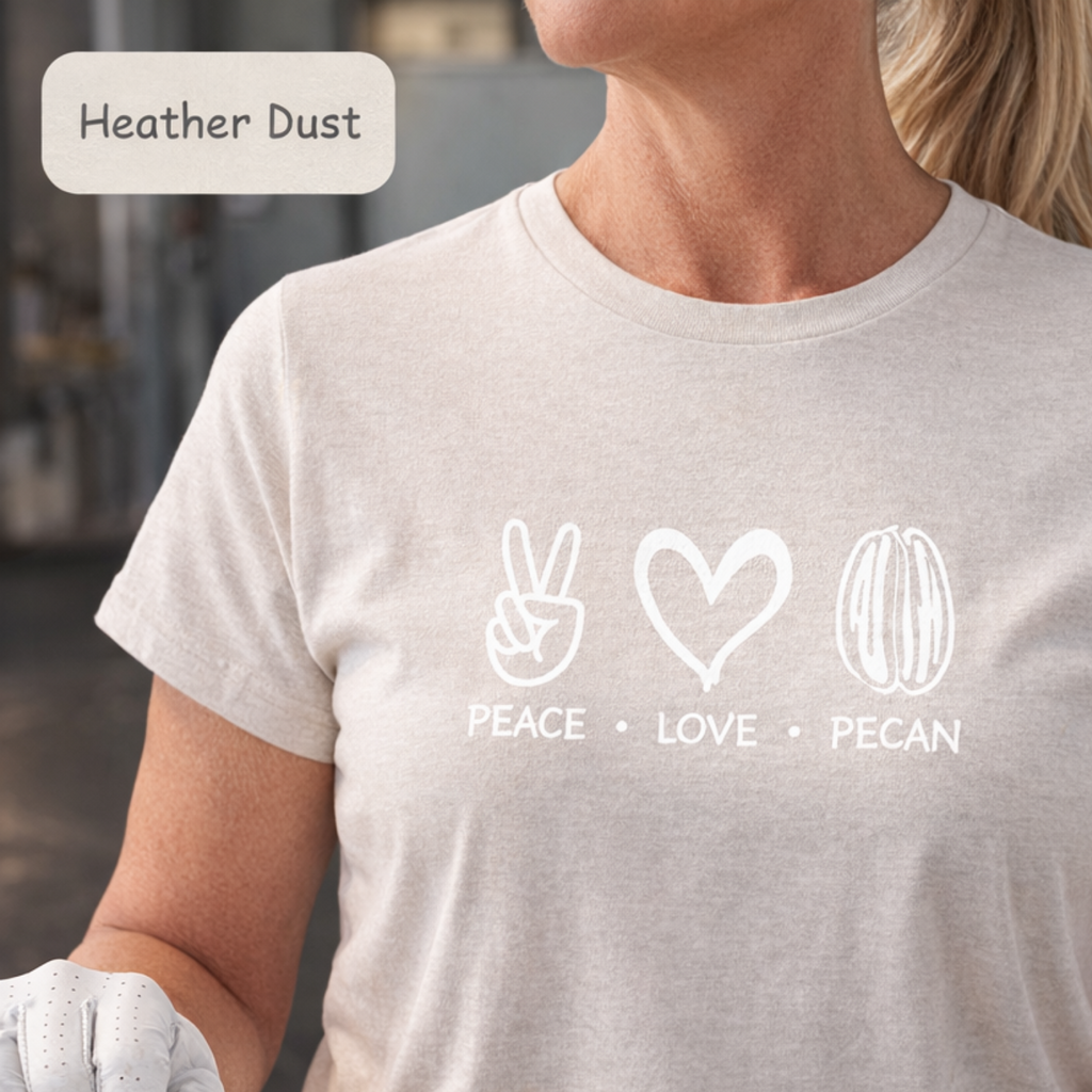 A woman wearing a light heather dust short-sleeve t-shirt with a white graphic print of peace, love, and pecan symbols with the text 'PEACE • LOVE • PECAN'.