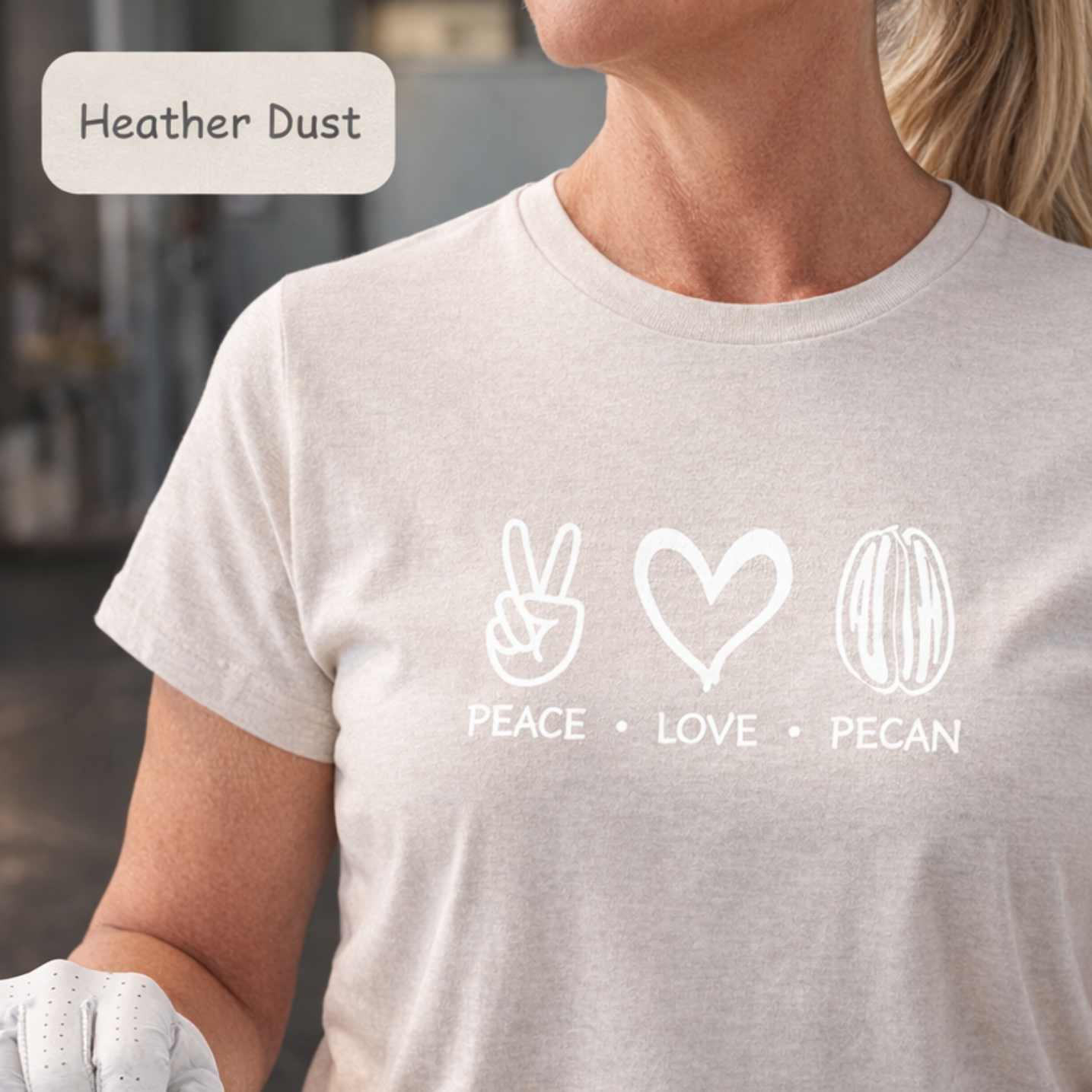 A woman wearing a light heather dust short-sleeve t-shirt with a white graphic print of peace, love, and pecan symbols with the text 'PEACE • LOVE • PECAN'.