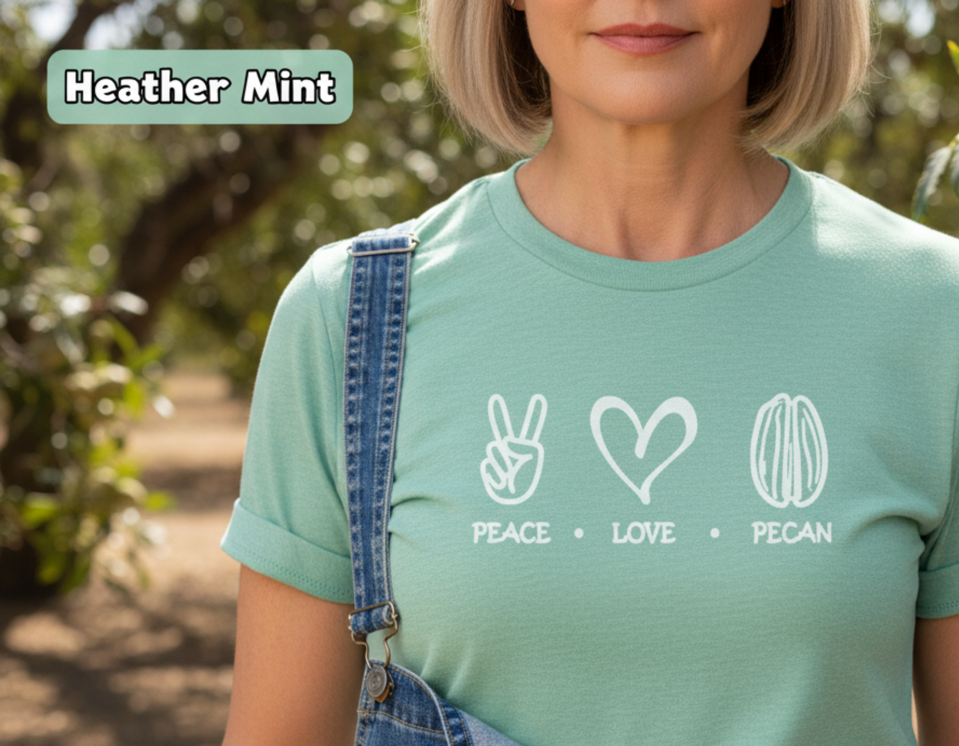 A woman outdoors wearing a heather mint green t-shirt with a white graphic of peace, love, and pecan symbols, paired with denim overalls. A 'Heather Mint' color label is in the top-left corner.