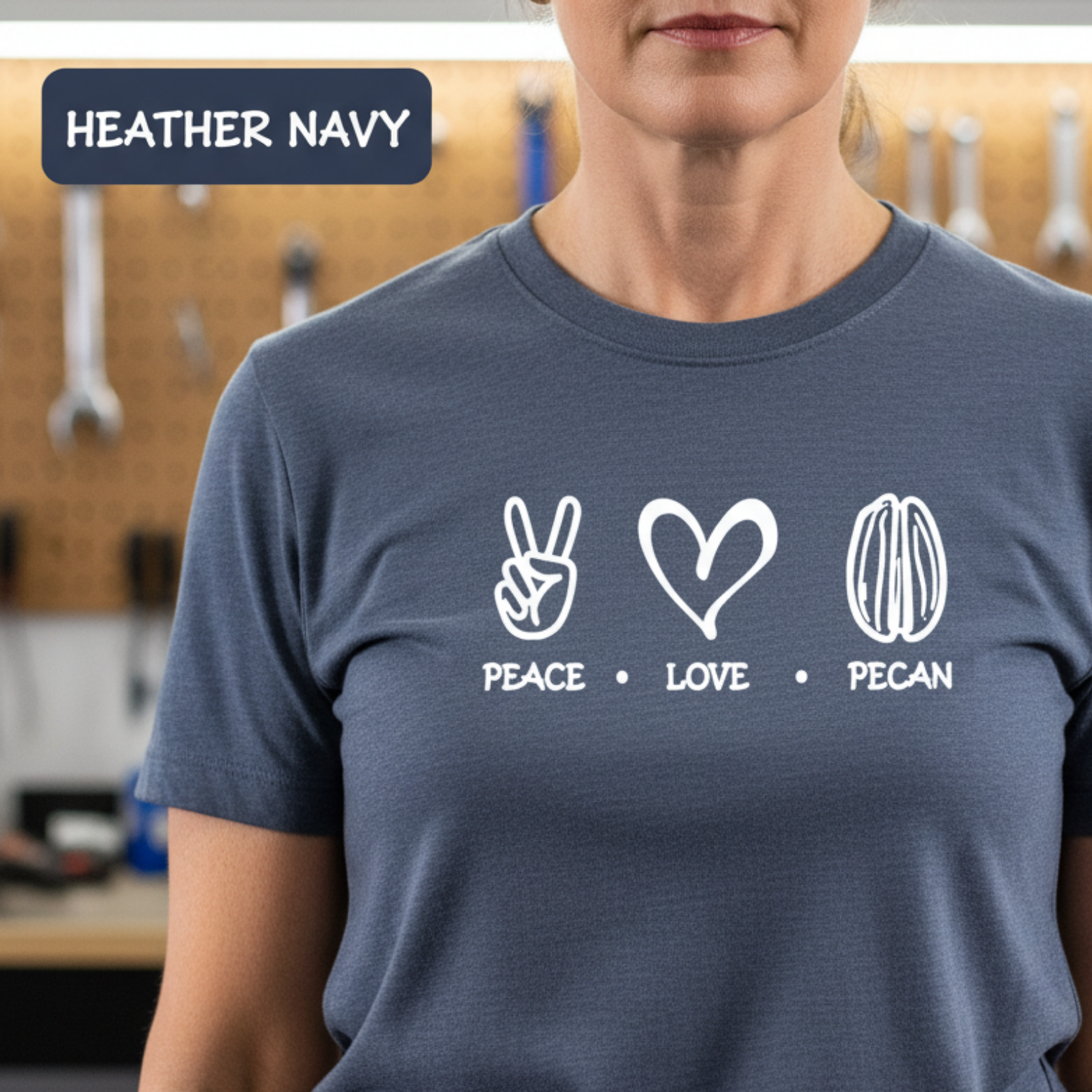 A person wearing a heather navy blue t-shirt with a white graphic print of peace, love, and pecan symbols with the words 'PEACE • LOVE • PECAN' underneath. The background shows a workshop with tools on a pegboard, and a label in the top left corner reads 'HEATHER NAVY'.