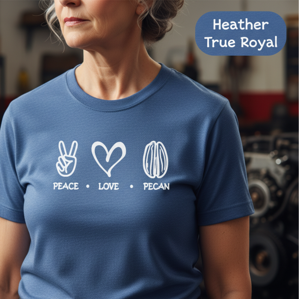 A woman wearing a heather blue short-sleeve t-shirt with a white graphic print of peace, love, and pecan symbols with the words 'PEACE • LOVE • PECAN' underneath. The background is a blurred workshop environment, and a blue label in the top right reads 'Heather True Royal'.