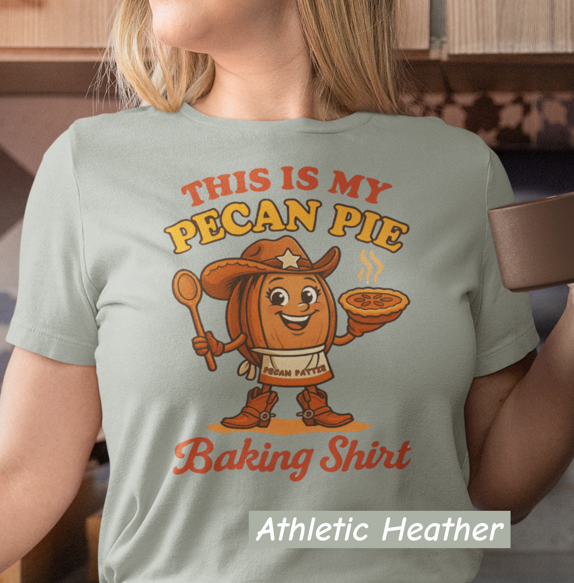 Ladies tee featuring cartoon Pecan Pattie wearing apron and hat, holding a pie and spoon, with the phrase ‘This Is My Pecan Pie Baking Shirt’ above and below her.”