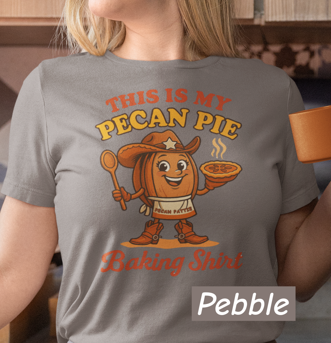 Ladies tee featuring cartoon Pecan Pattie wearing apron and hat, holding a pie and spoon, with the phrase ‘This Is My Pecan Pie Baking Shirt’ above and below her.”