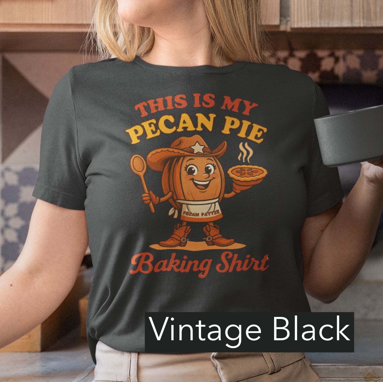 Ladies tee featuring cartoon Pecan Pattie wearing apron and hat, holding a pie and spoon, with the phrase ‘This Is My Pecan Pie Baking Shirt’ above and below her.”