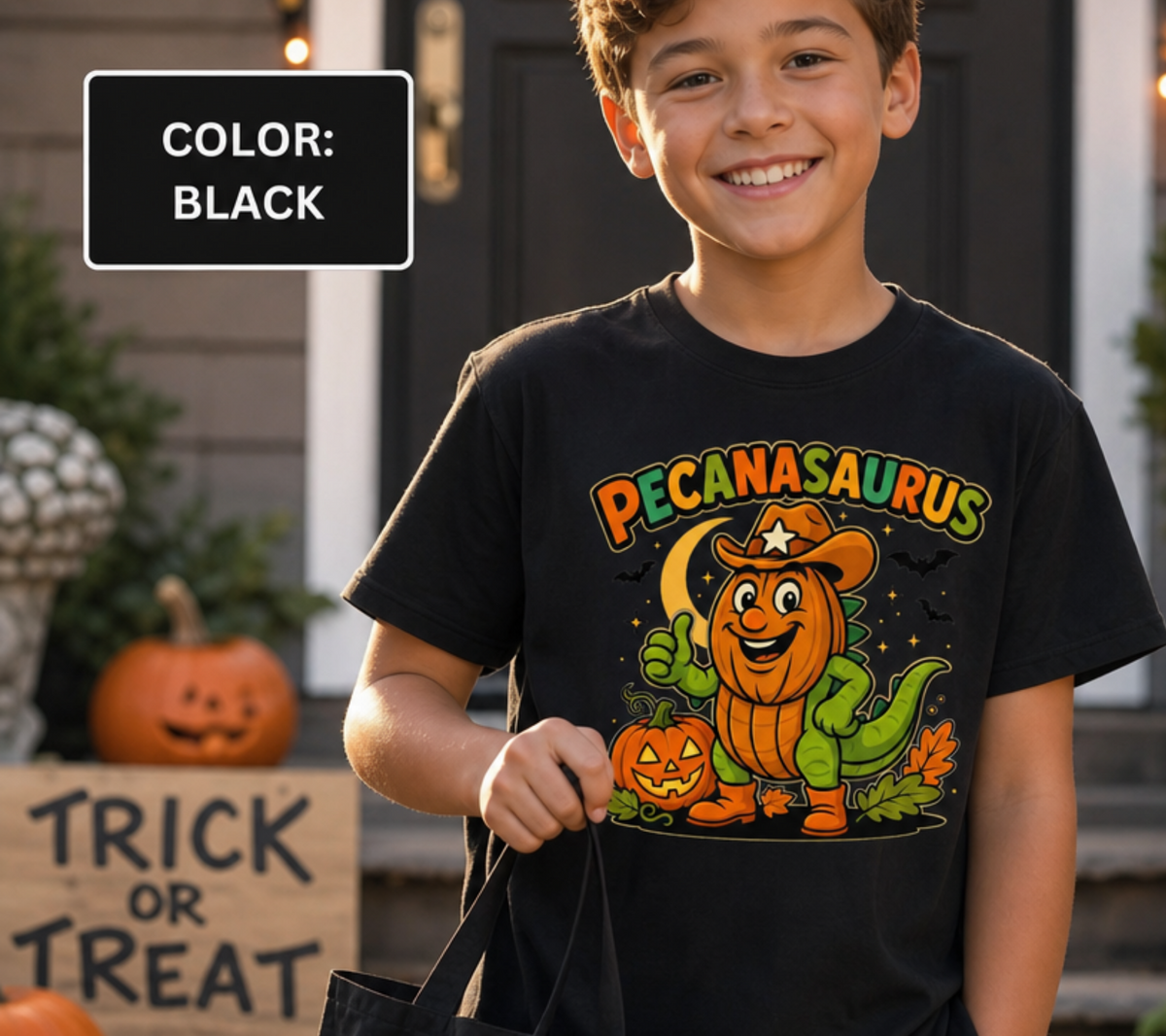 Black t-shirt with a Halloween graphic featuring the word "PECANASAURUS" above a green dinosaur wearing a pumpkin head, cowboy hat, and boots, set against a dark night background with a crescent moon, bats, and jack-o'-lanterns.