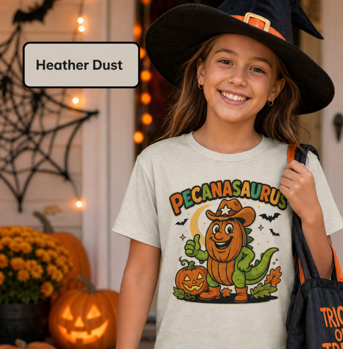 A smiling girl in a witch hat wears a light heather dust graphic T-shirt with a colorful 'Pecanasaurus' Halloween print, standing in a decorated porch setting with pumpkins and string lights. A label box reads 'Heather Dust'.