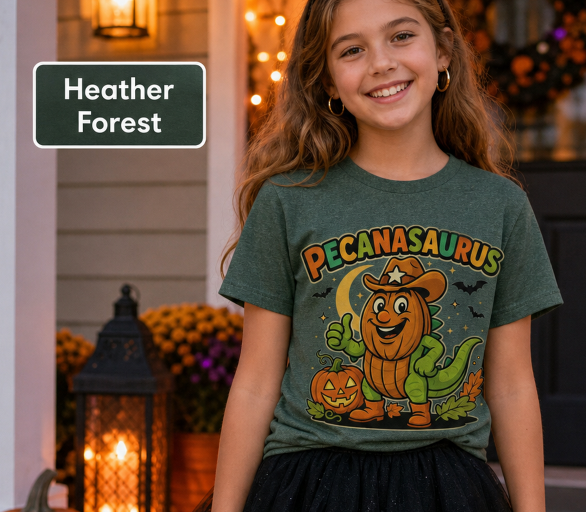 A smiling girl stands outdoors in front of a decorated front porch wearing a heather forest green graphic T-shirt. The shirt has a large Halloween-style print reading “PECANASaurus” with a cartoon pumpkin dinosaur, jack-o'-lantern, moon, and bats.