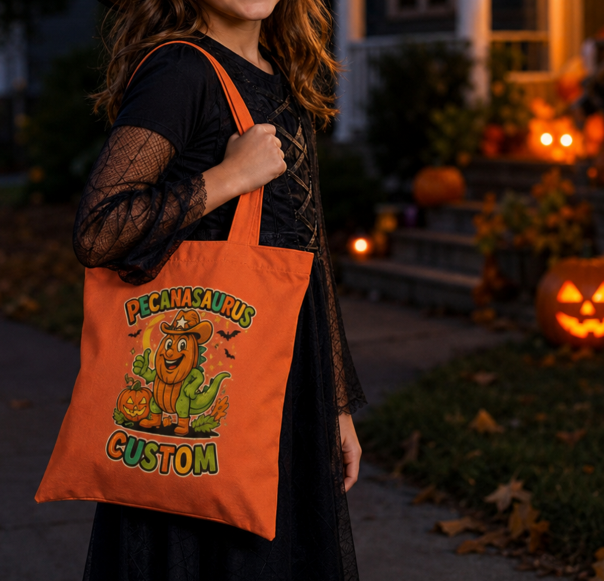 A person in a black lace dress holds an orange tote bag printed with a cartoon cactus wearing a sombrero and the words 'PECANSAURUS CUSTOM' in a Halloween yard at dusk.