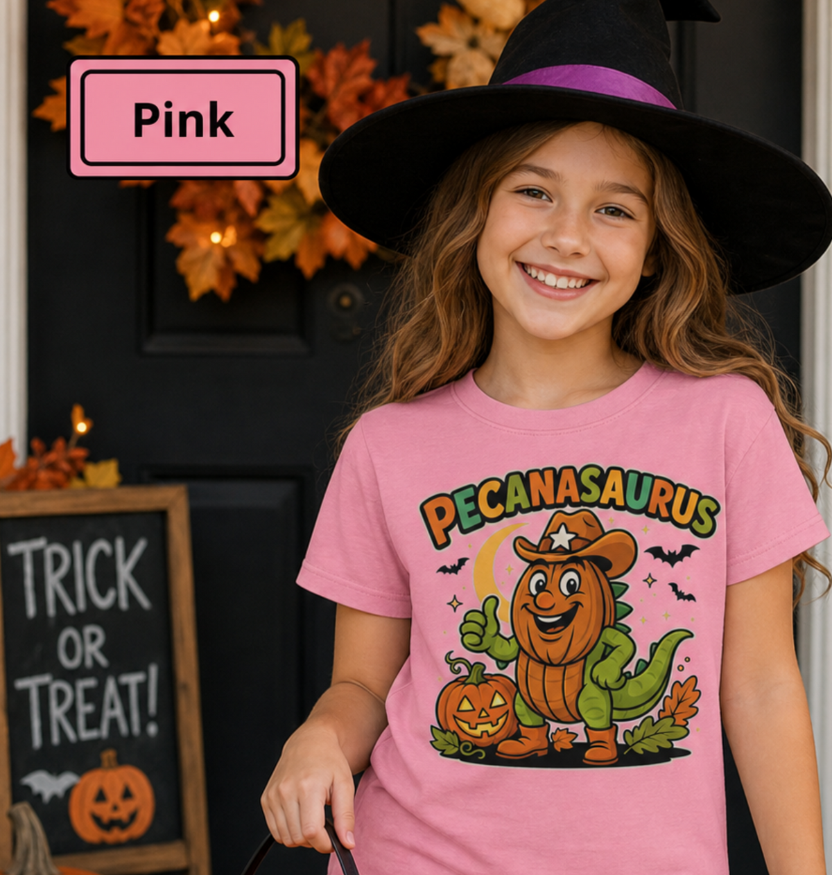 Pink t-shirt with a Halloween graphic featuring the word "PECANASAURUS" above a green dinosaur wearing a pumpkin head, cowboy hat, and boots, set against a dark night background with a crescent moon, bats, and jack-o'-lanterns.