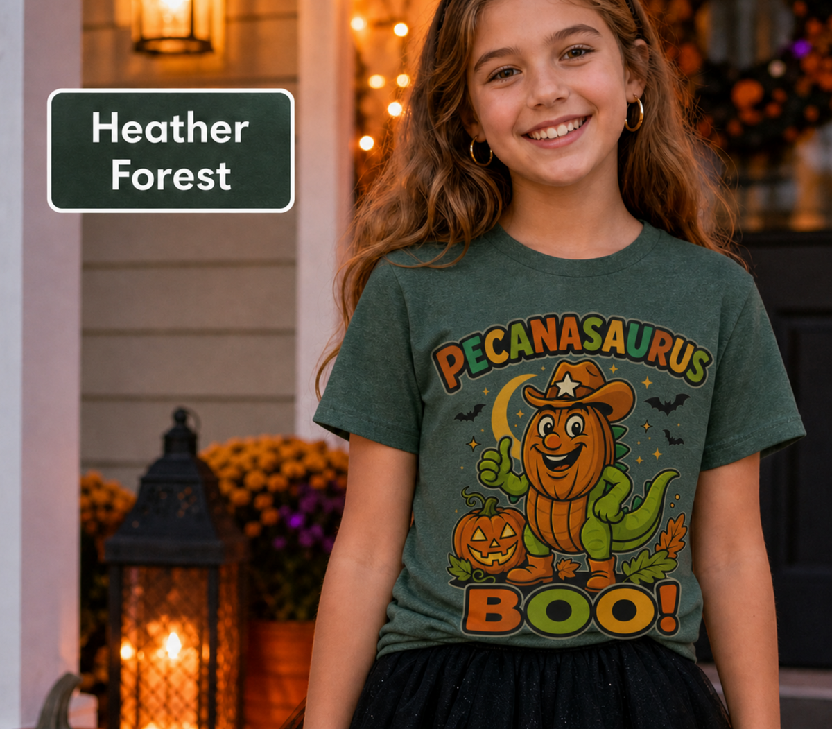 A smiling girl wearing a heather forest green graphic T-shirt with a cartoon pecan dinosaur Halloween design and the words 'Pecanaurus Boo!'. A dark green label in the top left reads 'Heather Forest'.
