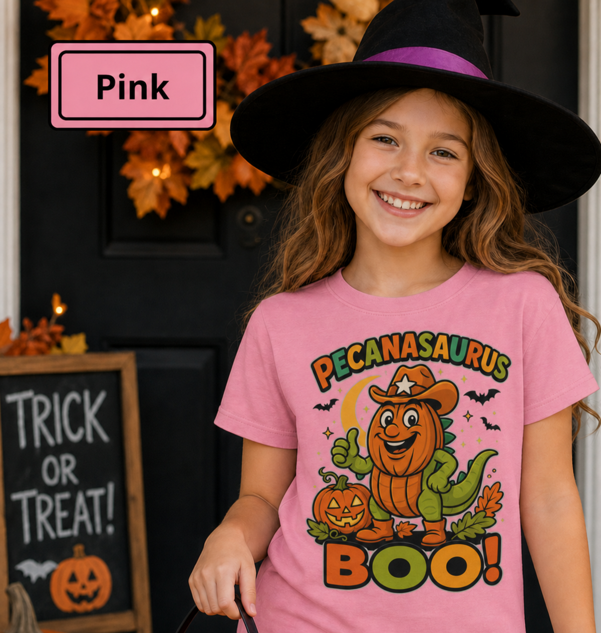 A young girl wearing a black witch hat with a purple band stands in front of a dark door decorated with autumn leaves. She is wearing a pink graphic T-shirt with a cartoon pumpkin dinosaur character and the words 'Pecanasaurus Boo!'.